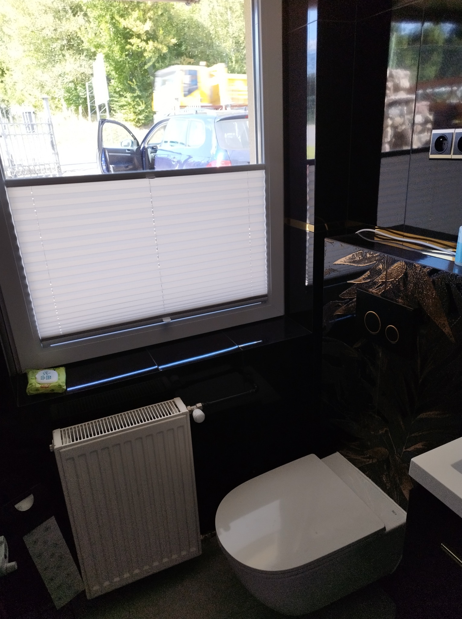 Nowoczesna łazienka z czarnymi płytkami imitującymi marmur, białą ceramiką i grzejnikiem. W oknie widoczny samochód i zieleń. Minimalistyczny design, eleganckie wykończenie.