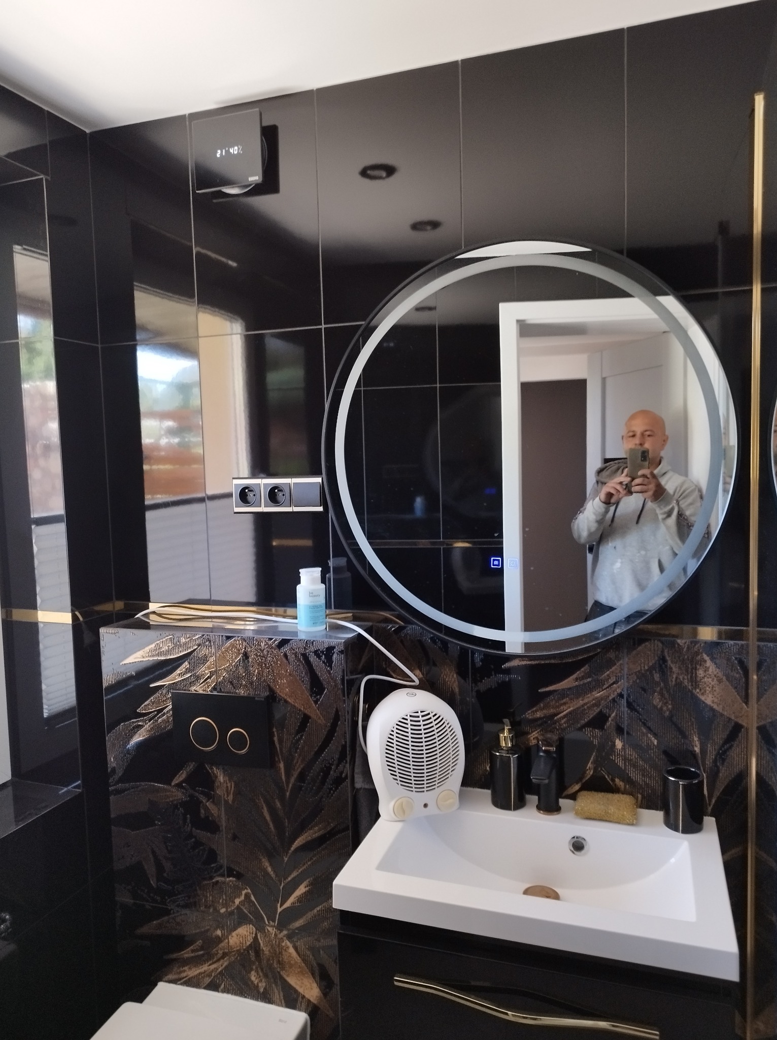 Wykończona łazienka w czerni i złocie: czarne płytki, umywalka z szafką, okrągłe lustro z podświetleniem, dekoracyjne wzory na ścianach. Widoczne gniazdka i wentylator.