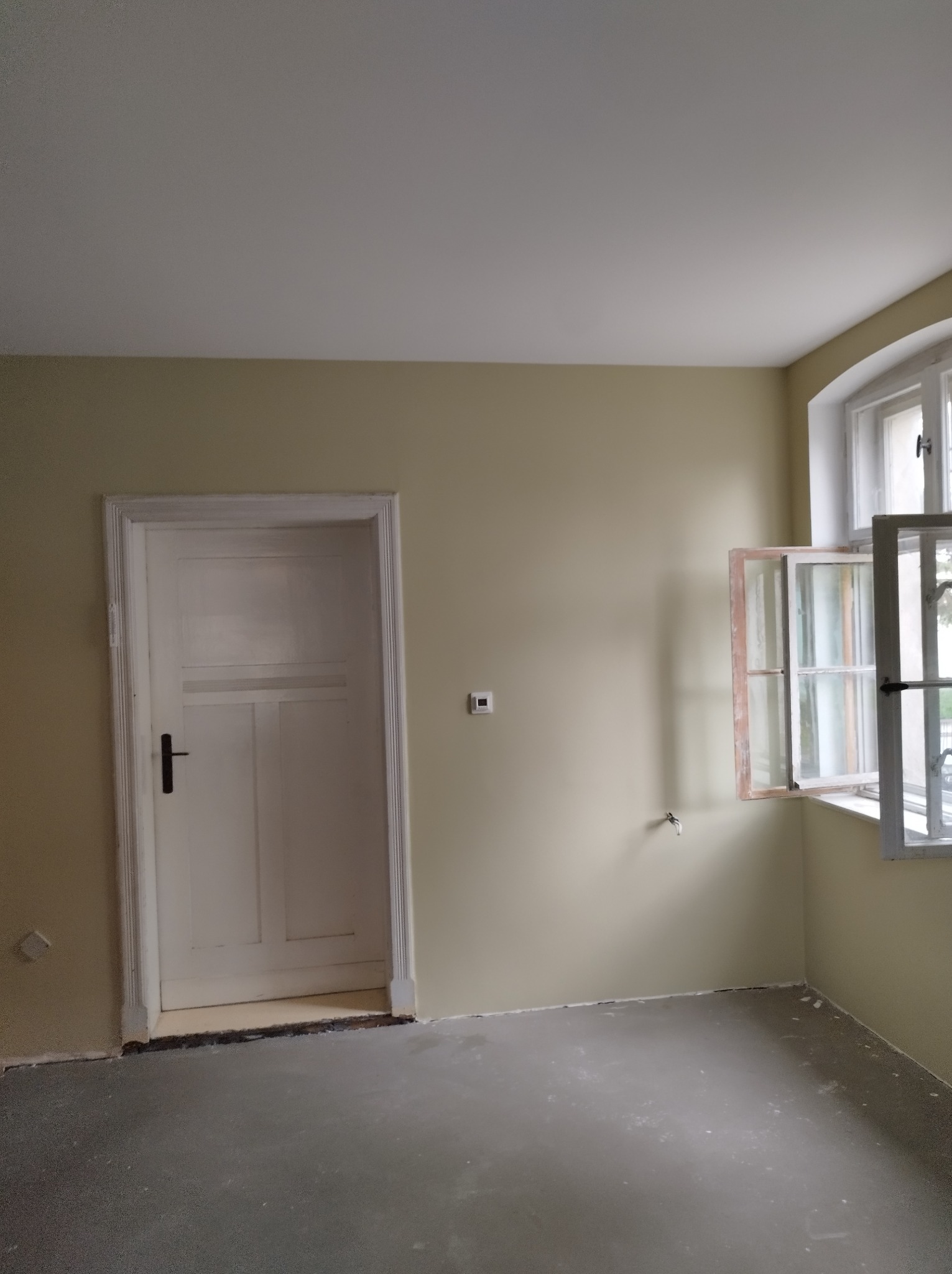 Pomieszczenie po malowaniu ścian na beżowo, z białymi drzwiami i oknem. Widoczna świeżo pomalowana podłoga. Otwarta okiennica.