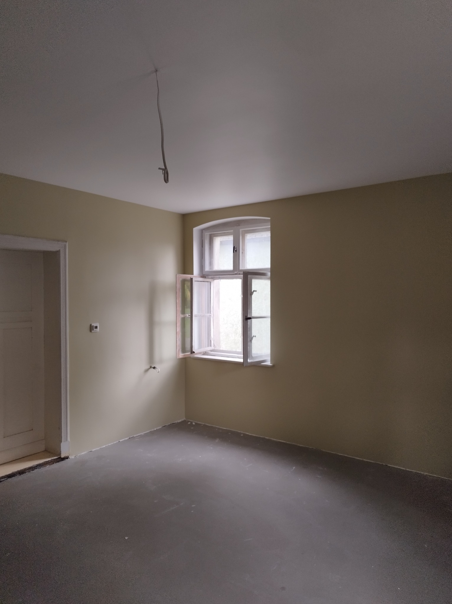 Pomalowane na beżowo wnętrze pokoju z otwartym oknem i szarą podłogą. Widoczne kable zwisające z sufitu. Świeżo odnowione ściany i podłoga.