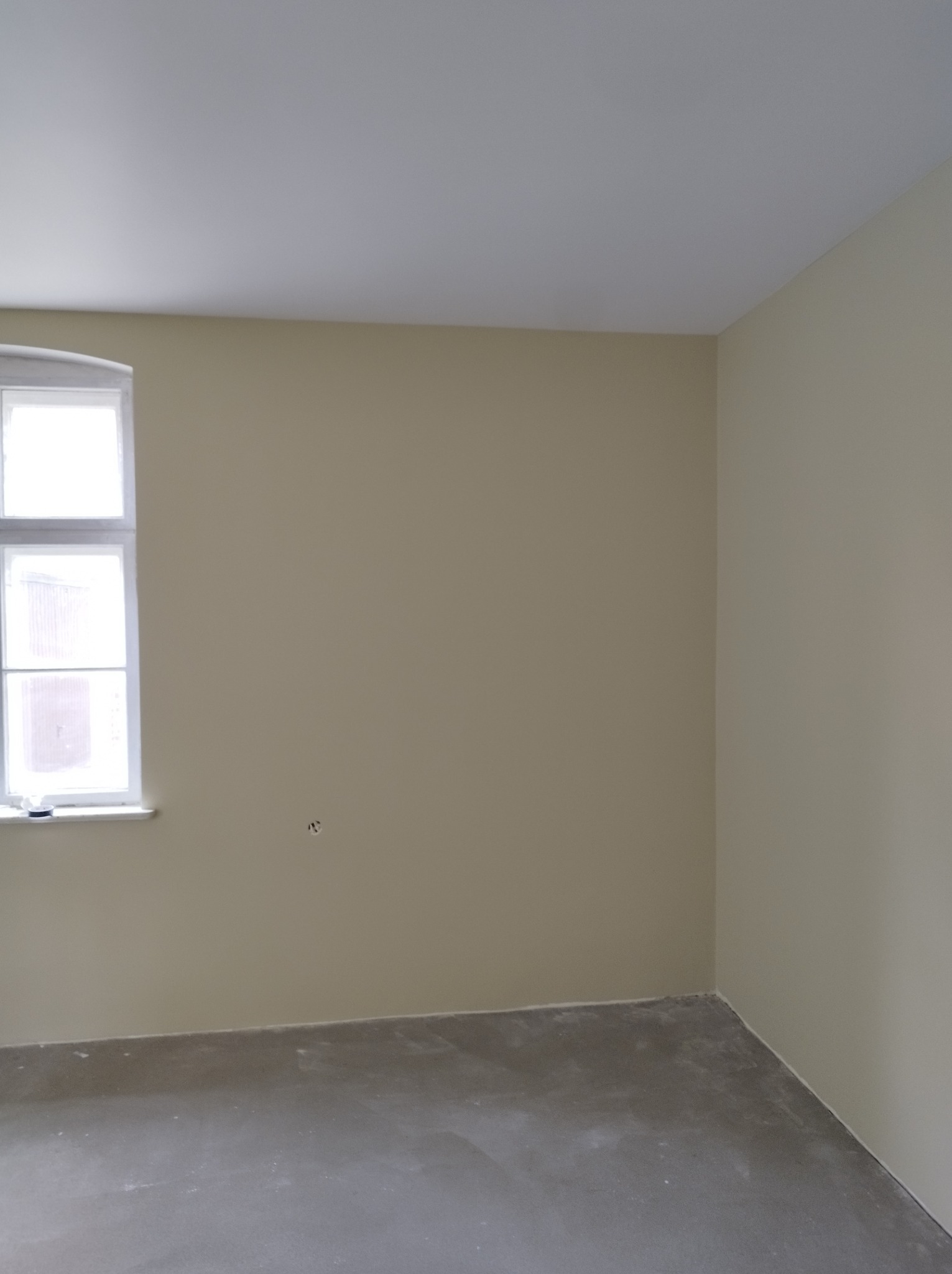 Pomalowane na beżowo wnętrze pokoju z oknem. Na ścianie widoczny otwór po gniazdku. Podłoga surowa, przygotowana do dalszych prac wykończeniowych.