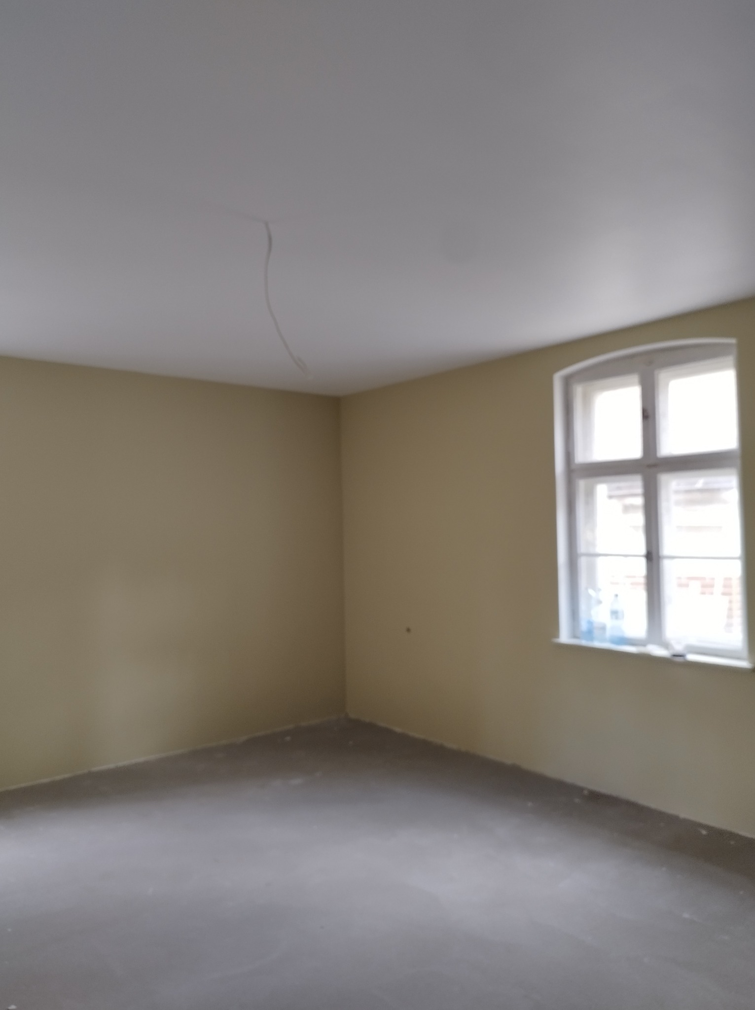 Pomalowane na beżowo wnętrze pokoju z oknem. Widoczne ściany, sufit z kablem i szara podłoga. Świeżo po malowaniu, gotowe do dalszych prac wykończeniowych.