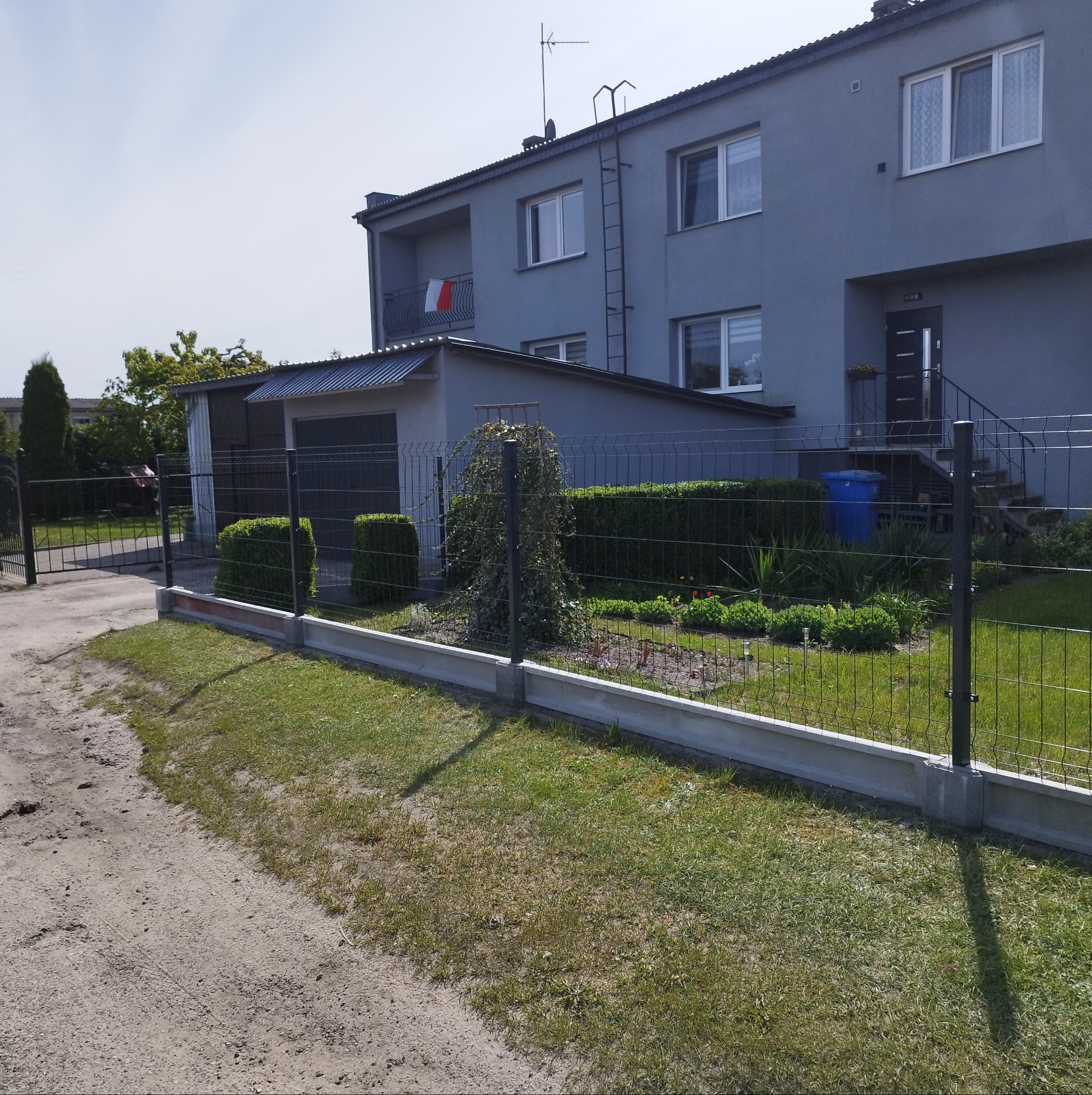 Szara elewacja dwukondygnacyjnego domu z balkonem, widoczna zza nowego, metalowego ogrodzenia z betonową podmurówką i zadbanym trawnikiem. Słoneczny dzień.
