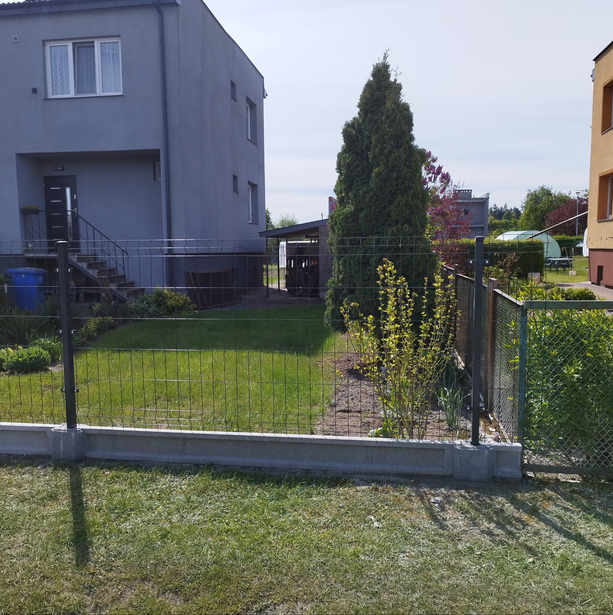 Nowoczesne ogrodzenie panelowe z podmurówką betonową, otaczające zielony ogród z drzewami i krzewami, na tle szarego domu dwukondygnacyjnego w słoneczny dzień.