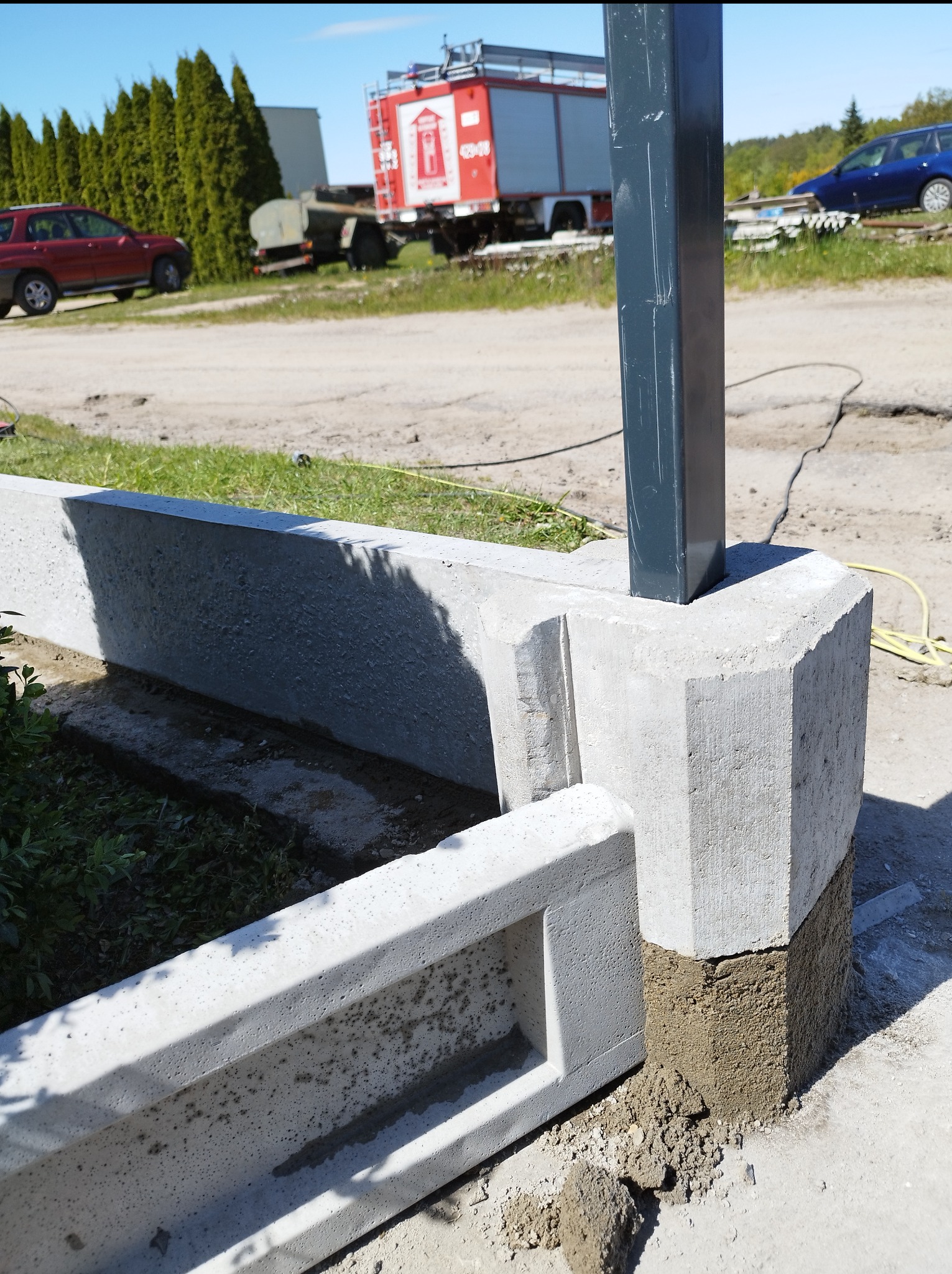 Montaż betonowych obrzeży trawnikowych z metalowym słupkiem. Widoczne niedociągnięcia w fugowaniu. W tle wóz strażacki i zieleń.
