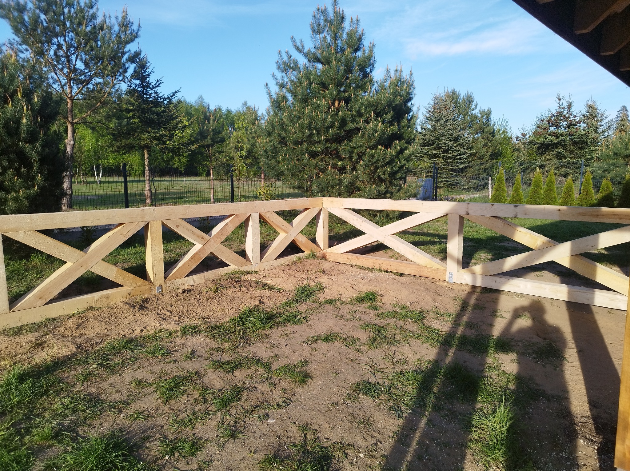 Drewniane ogrodzenie w stylu rustykalnym, z krzyżowymi belkami, otoczone zielenią i drzewami iglastymi. Widok na błękitne niebo w słoneczny dzień.