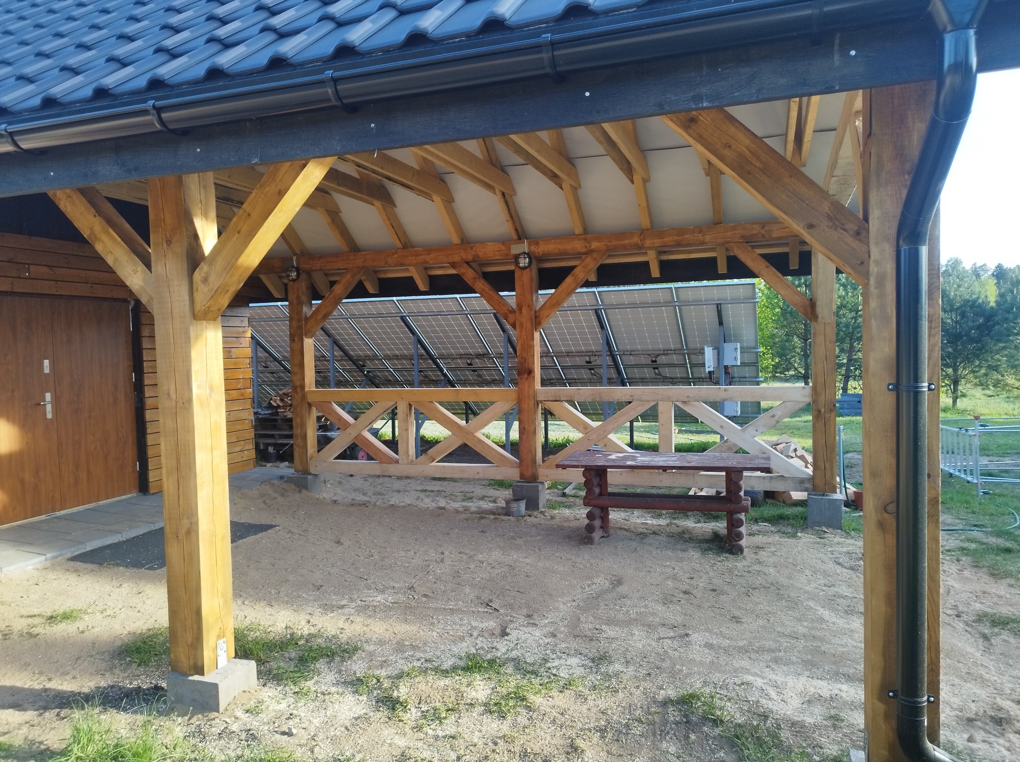 Drewniana altana z widocznymi panelami słonecznymi w tle. Konstrukcja z belek, zadaszenie z dachówki. Na ziemi piasek i mały drewniany stół.