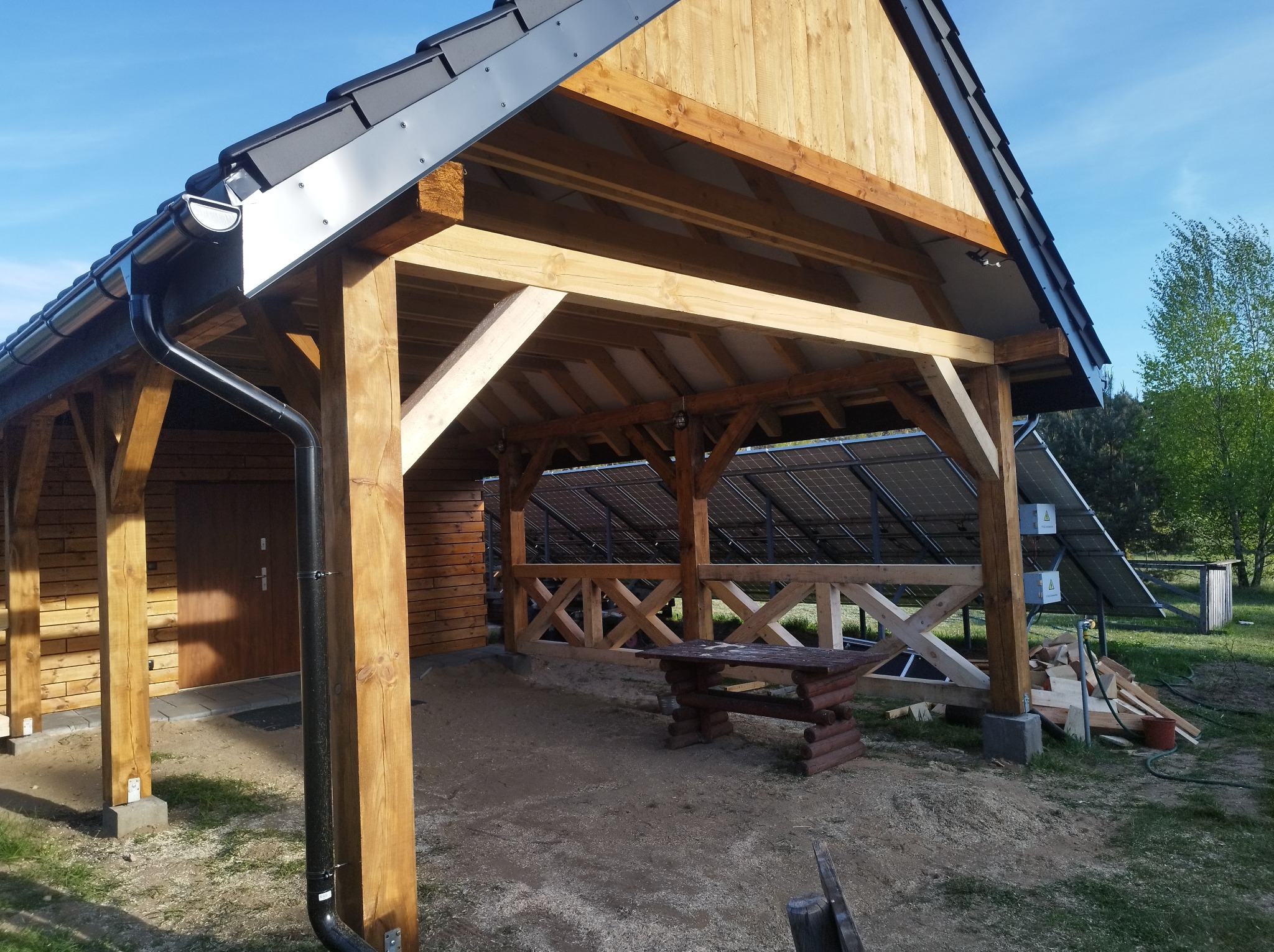 Drewniana konstrukcja z zadaszeniem i rynną, widoczne panele słoneczne w tle. Elementy drewniane w naturalnym kolorze, czarna rynna. Na ziemi wióry i resztki materiałów.