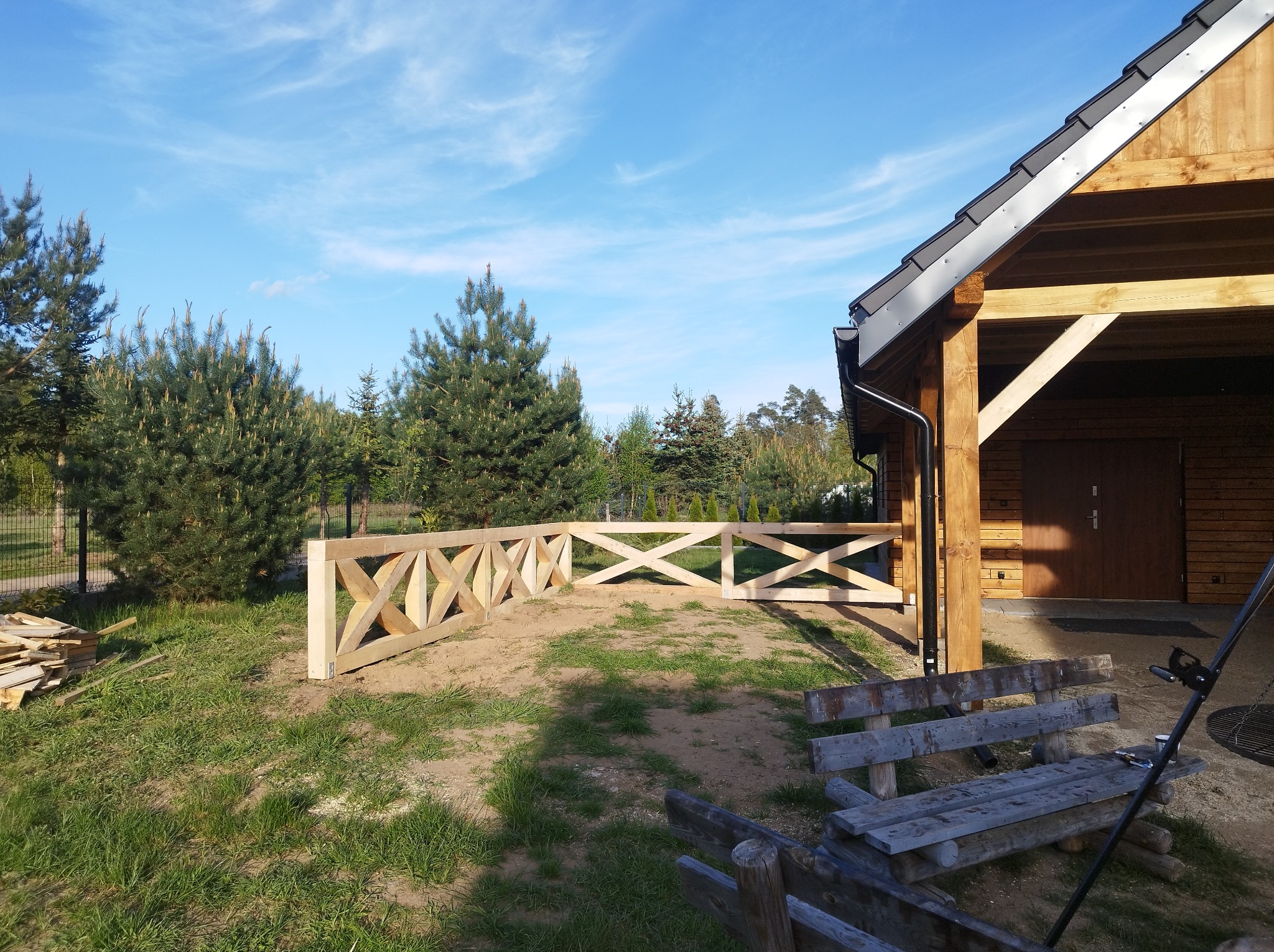 Drewniana, surowa balustrada z krzyżowym wzorem, otaczająca teren przy drewnianym budynku w stylu rustykalnym, na tle zieleni i błękitnego nieba.