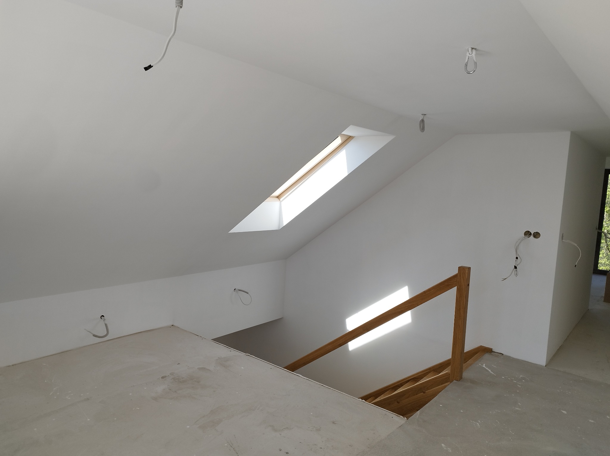 Poddasze w trakcie wykańczania: białe ściany, okno dachowe wpuszczające światło, drewniana balustrada schodów, widoczne kable i surowa podłoga.