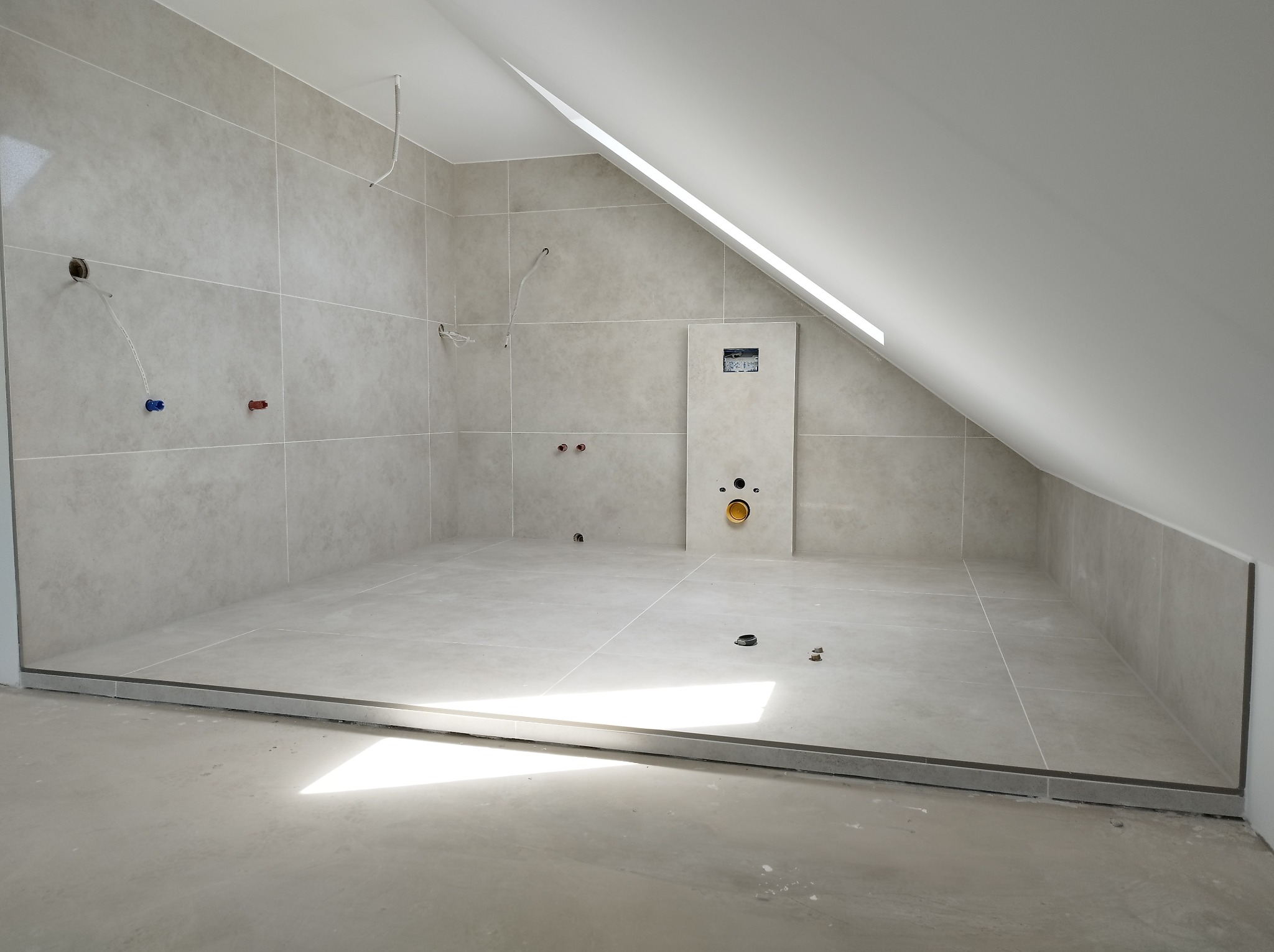 Wykończona łazienka na poddaszu z beżowymi płytkami, widoczne przyłącza wodne i kanalizacyjne. Ułożona podłoga z odpływem liniowym. Skos dachu z oknem.