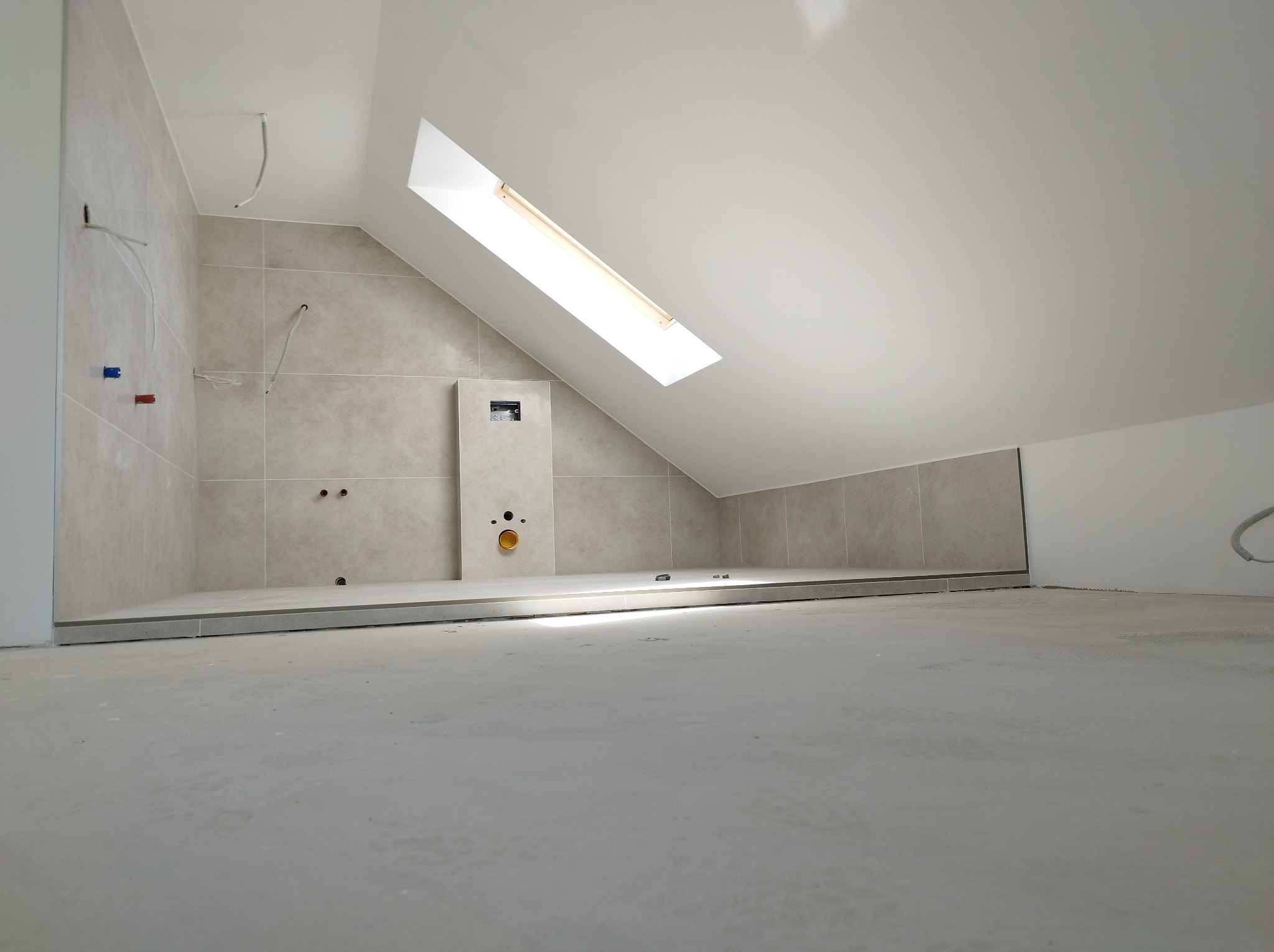 Wykończona łazienka na poddaszu z beżowymi płytkami, widoczne instalacje i okno dachowe. Minimalistyczny design, surowy beton na podłodze.