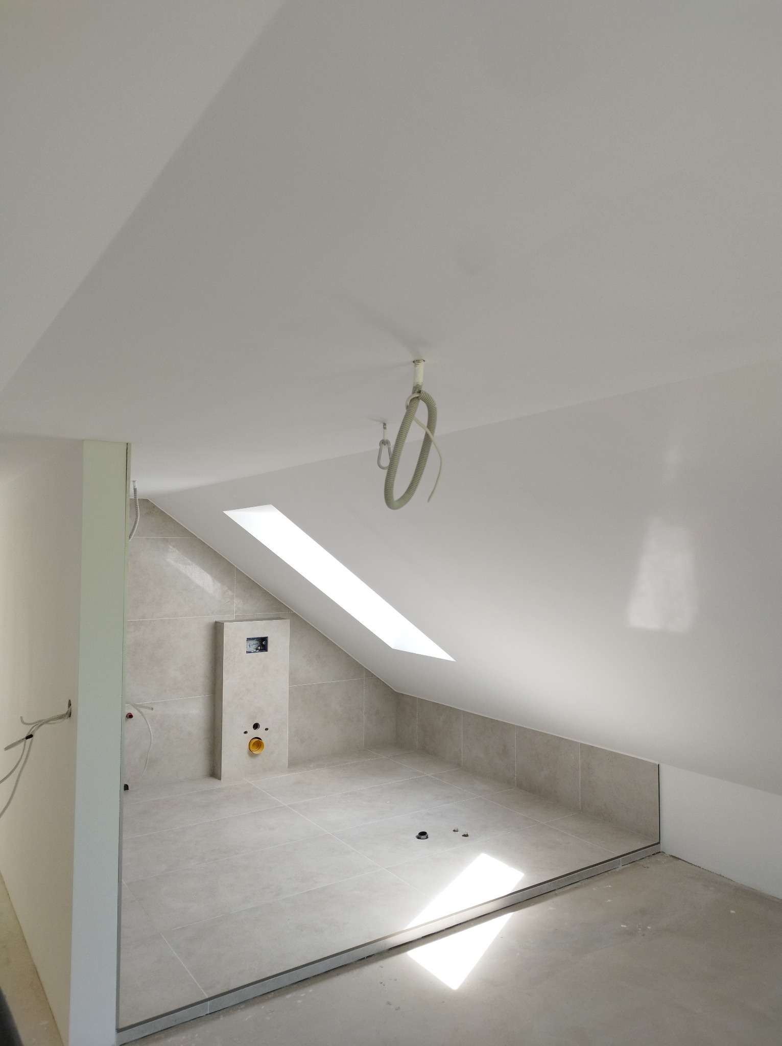 Wykończona łazienka na poddaszu z beżowymi płytkami, oknem dachowym i białym sufitem. Widoczne instalacje i fragment surowej podłogi. Minimalistyczny design.