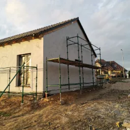 Dom jednorodzinny w trakcie prac elewacyjnych, z nałożonym szarym tynkiem i rozstawionym rusztowaniem, okno zabezpieczone folią, teren wokół domu częściowo uporządkowany.