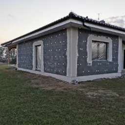 Dom jednorodzinny w trakcie ocieplania, widoczna szara warstwa izolacyjna z białymi kołkami, okna zabezpieczone folią, przygotowane do dalszych prac.