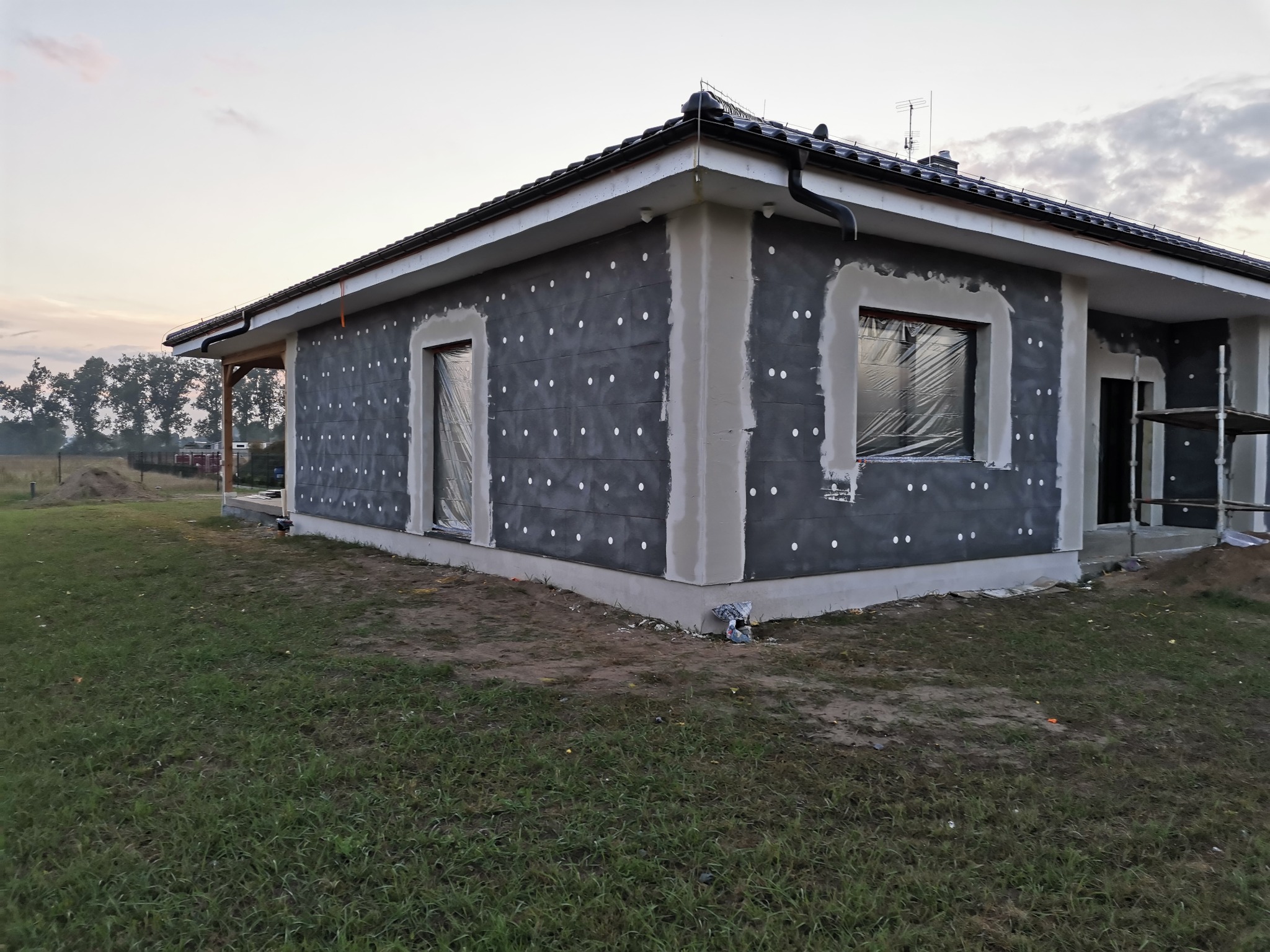 Dom jednorodzinny w trakcie ocieplania, widoczna szara warstwa izolacyjna z białymi kołkami, okna zabezpieczone folią, przygotowane do dalszych prac.