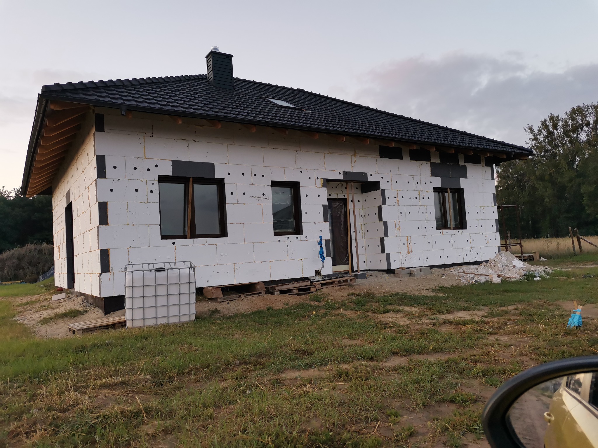 Dom jednorodzinny w trakcie ocieplania styropianem, widoczne czarne listwy montażowe, okna z brązowymi ramami, czarny dach z dachówką, zbiornik na wodę na palecie, trawa i drzewa w tle.
