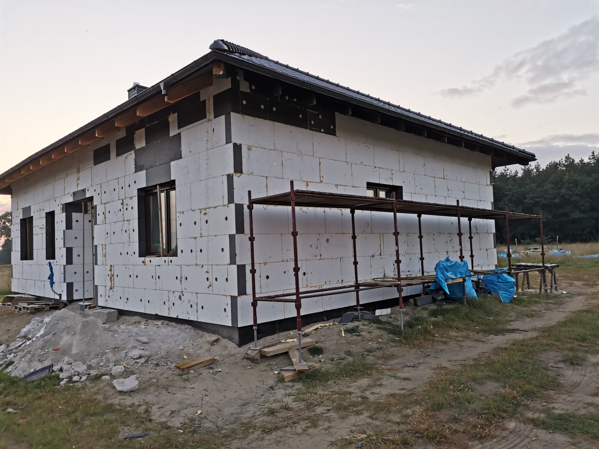 Niedokończony budynek jednorodzinny z widocznym ociepleniem ścian zewnętrznych, rusztowaniem i elementami czarnej izolacji, na tle drzew i nieba o zmierzchu.