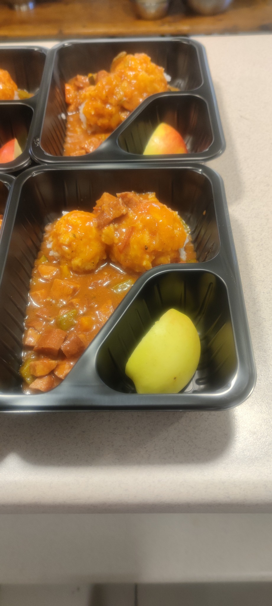 Dwa czarne pojemniki z jedzeniem na wynos: kulki ziemniaczane w sosie z kawałkami kiełbasy i zielonego pieprzu, obok plasterek jabłka.