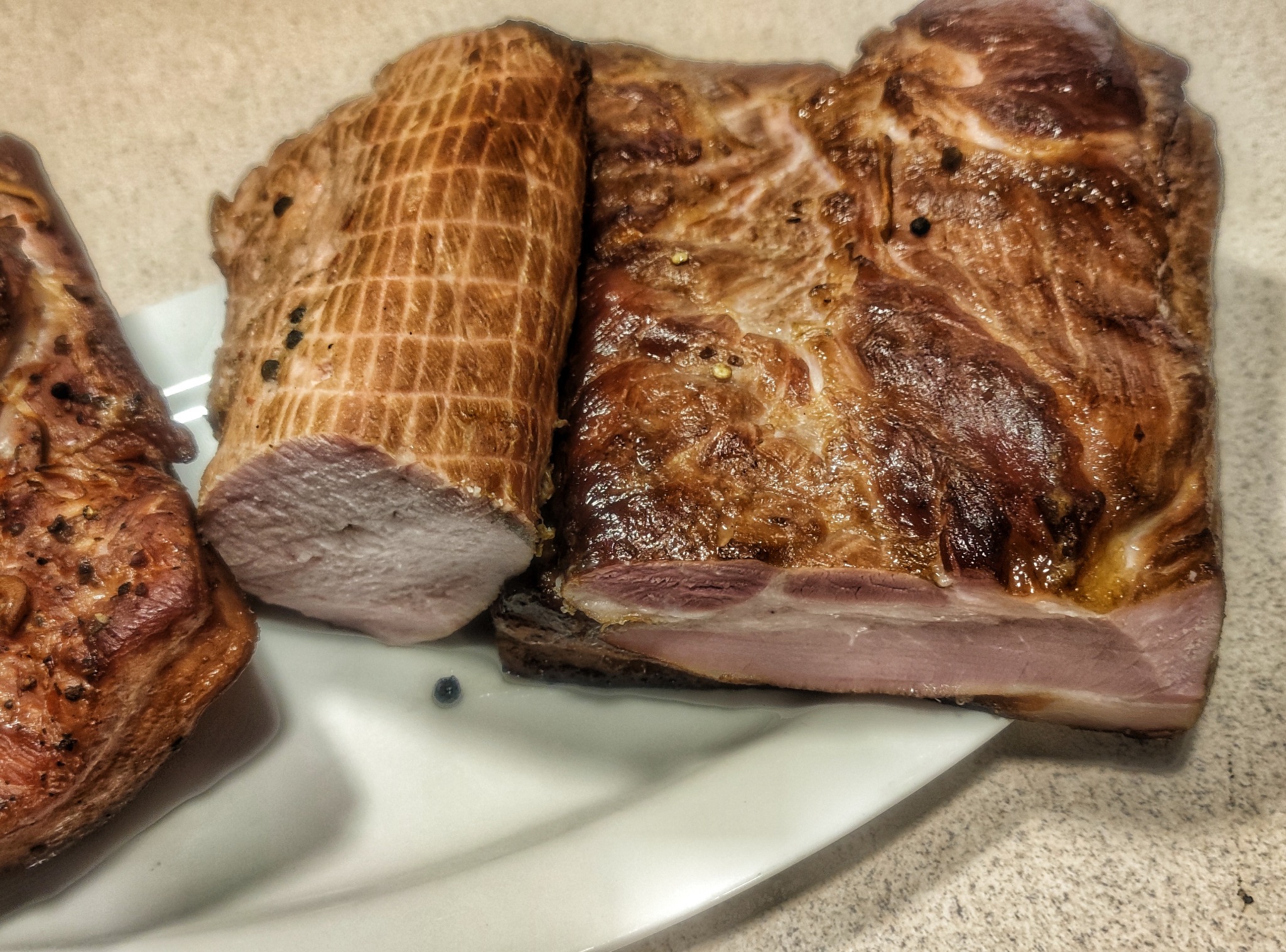 Plater z wędlinami: karkówka w siatce wędliniarskiej, pieczona szynka z widoczną warstwą tłuszczu i polędwica, posypane ziarnami pieprzu.