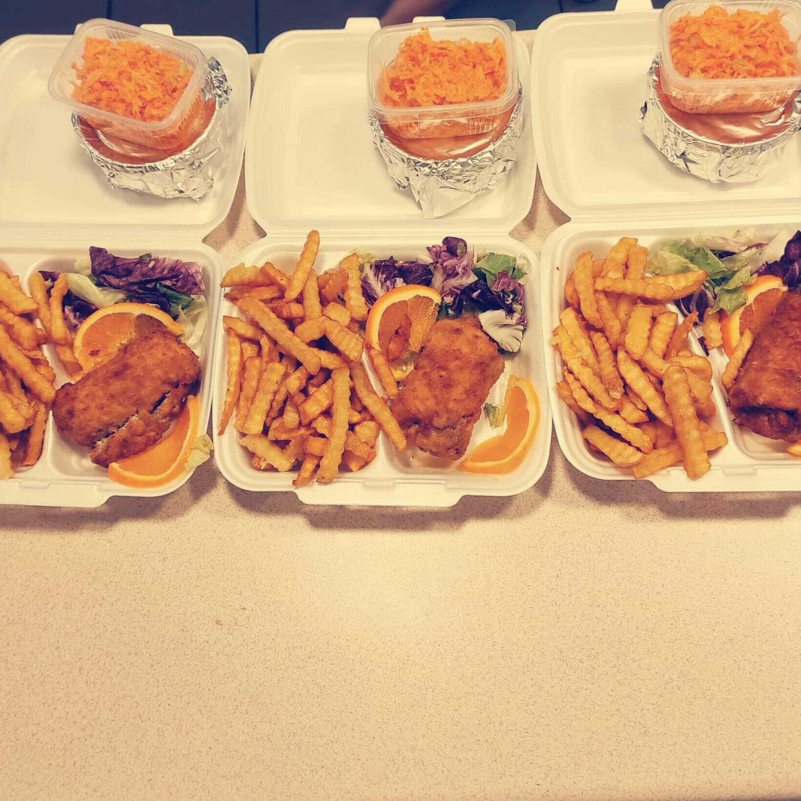 Trzy białe pudełka cateringowe z frytkami, kotletem w panierce, sałatką i plasterkiem pomarańczy; oddzielne pojemniki z surówką z marchewki owinięte w folię aluminiową.