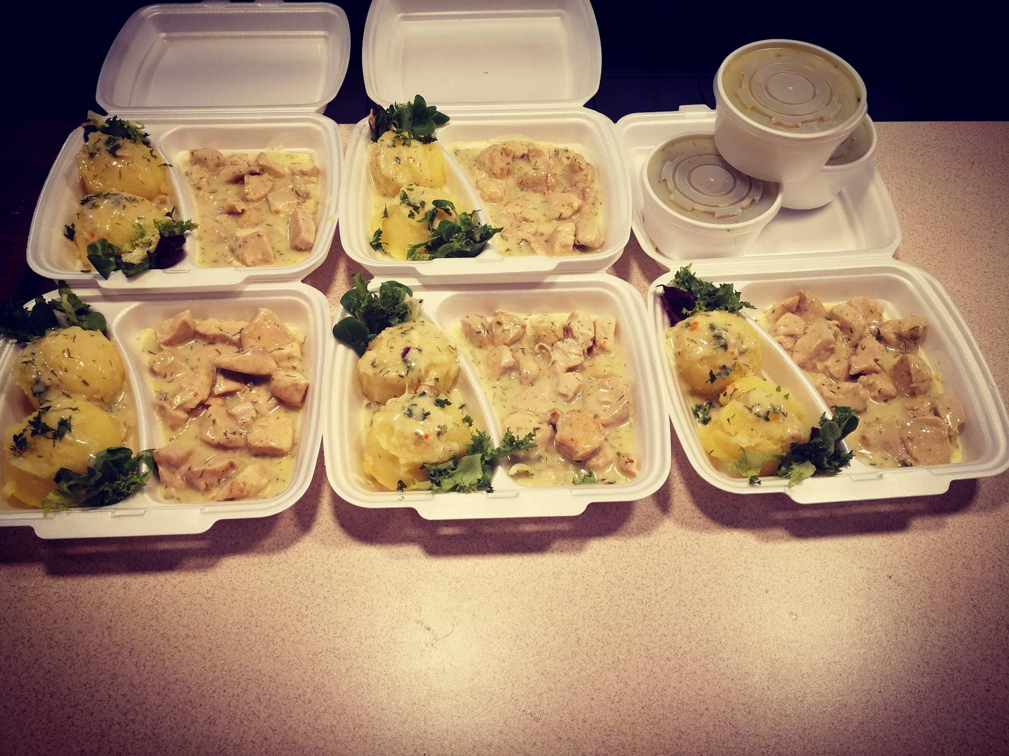 Porcje obiadowe w białych pojemnikach na wynos: ziemniaki z koperkiem i sałatą oraz kurczak w sosie śmietanowym, dodatkowo sos w okrągłych pojemnikach.