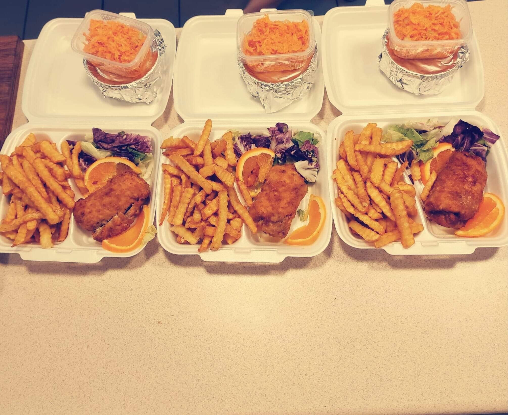 Trzy białe, styropianowe pojemniki z zestawami obiadowymi: smażona ryba, frytki, sałatka z pomarańczą i surówka z marchewki w osobnym pojemniku.