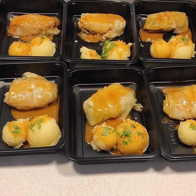 Porcje gołąbków z ziemniakami polane sosem, ułożone w czarnych, plastikowych pojemnikach, posypane świeżą natką pietruszki.