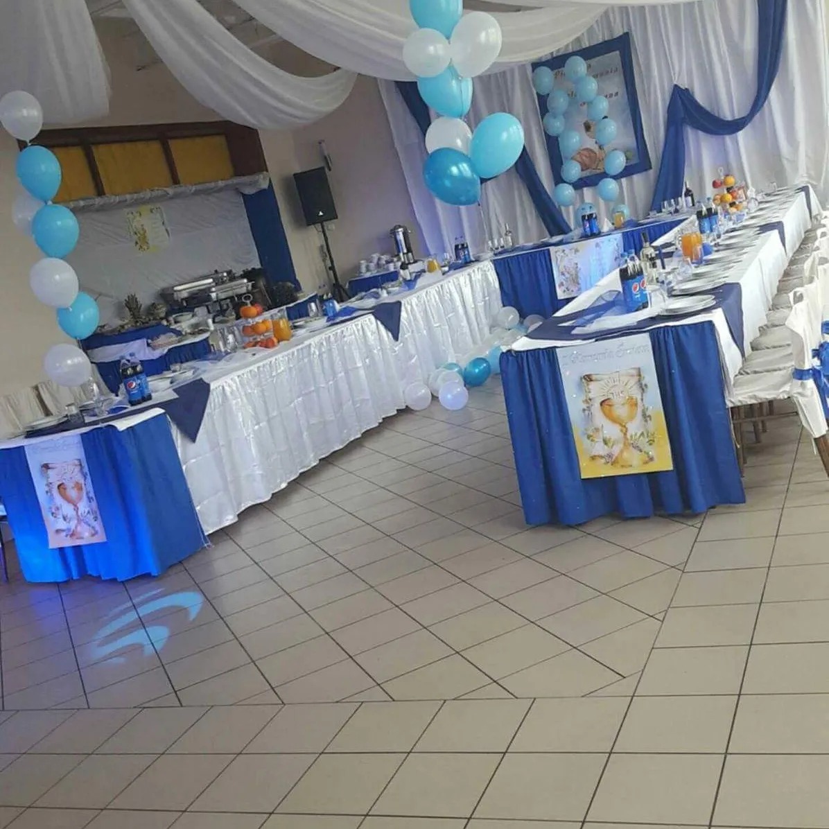 Udekorowana sala bankietowa z długimi stołami w białych obrusach z niebieskimi akcentami, niebieskie i białe balony, dekoracje z białego materiału pod sufitem, przygotowana na uroczystość komunijną.