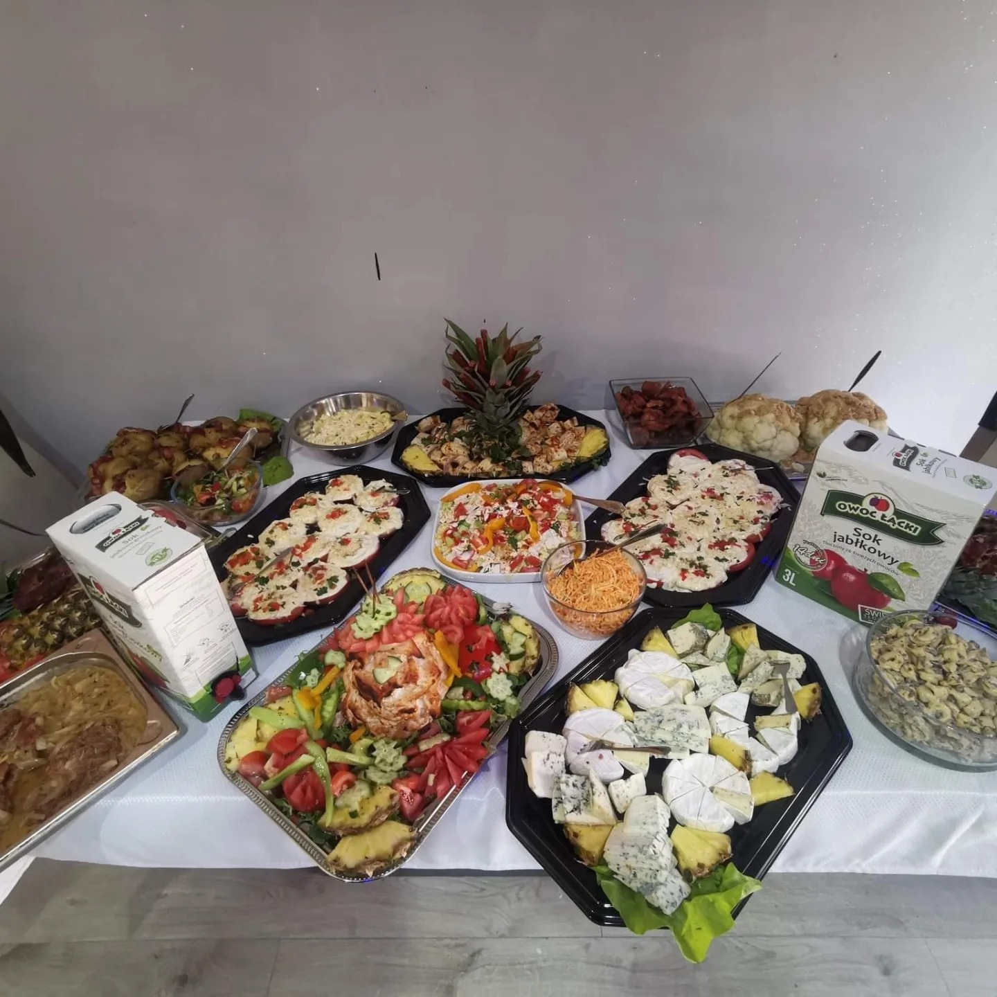 Stół zastawiony różnorodnymi potrawami cateringowymi: sałatki, sery pleśniowe z ananasem, koreczki, dania mięsne, sok jabłkowy 3L, całość na białym obrusie.