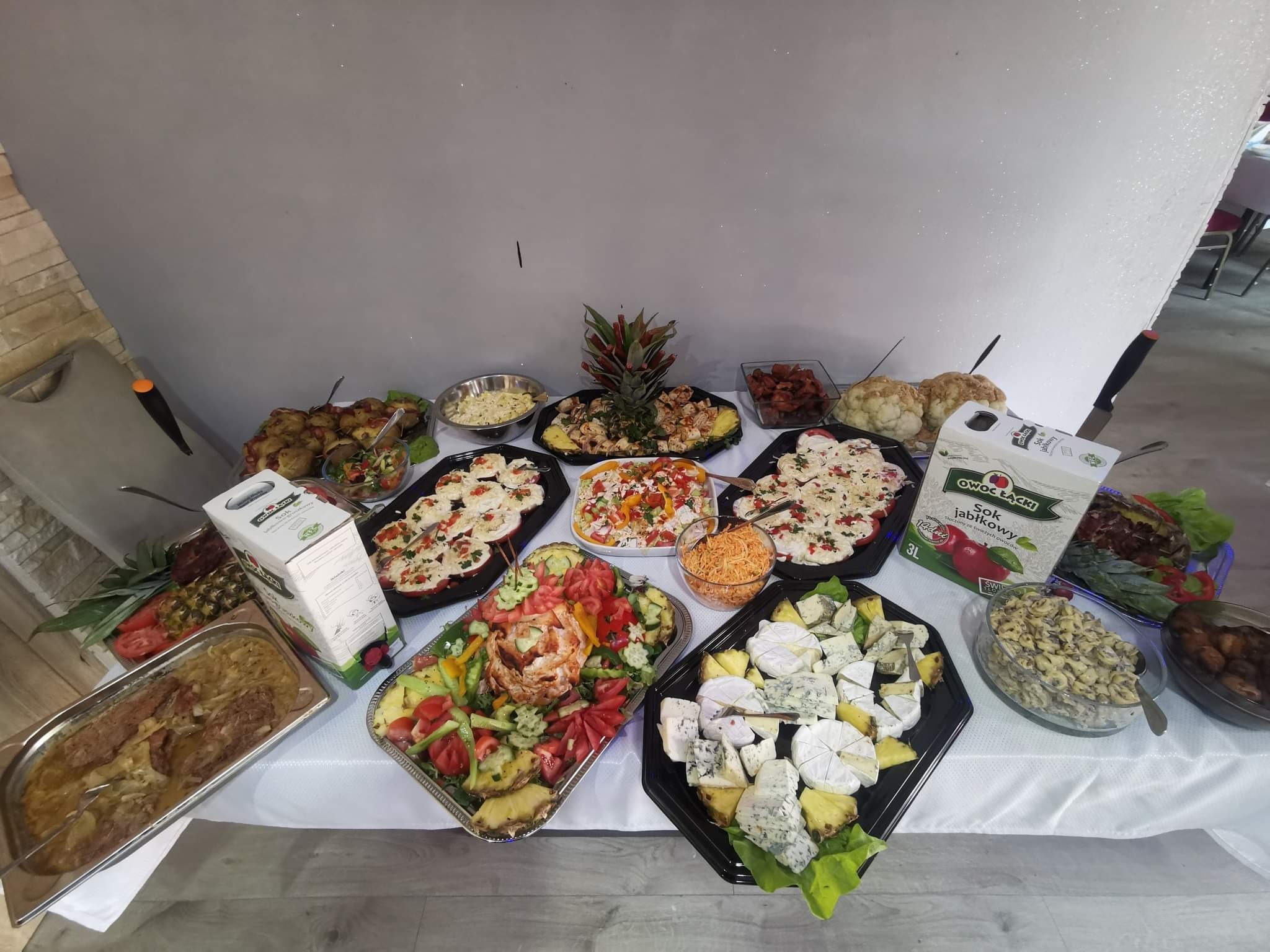 Stół z różnorodnymi potrawami: sałatki warzywne, sery pleśniowe z ananasem, dania mięsne w sosie, przekąski z pomidorami, całość dopełniona sokiem jabłkowym w kartonie i dekoracją z ananasa.