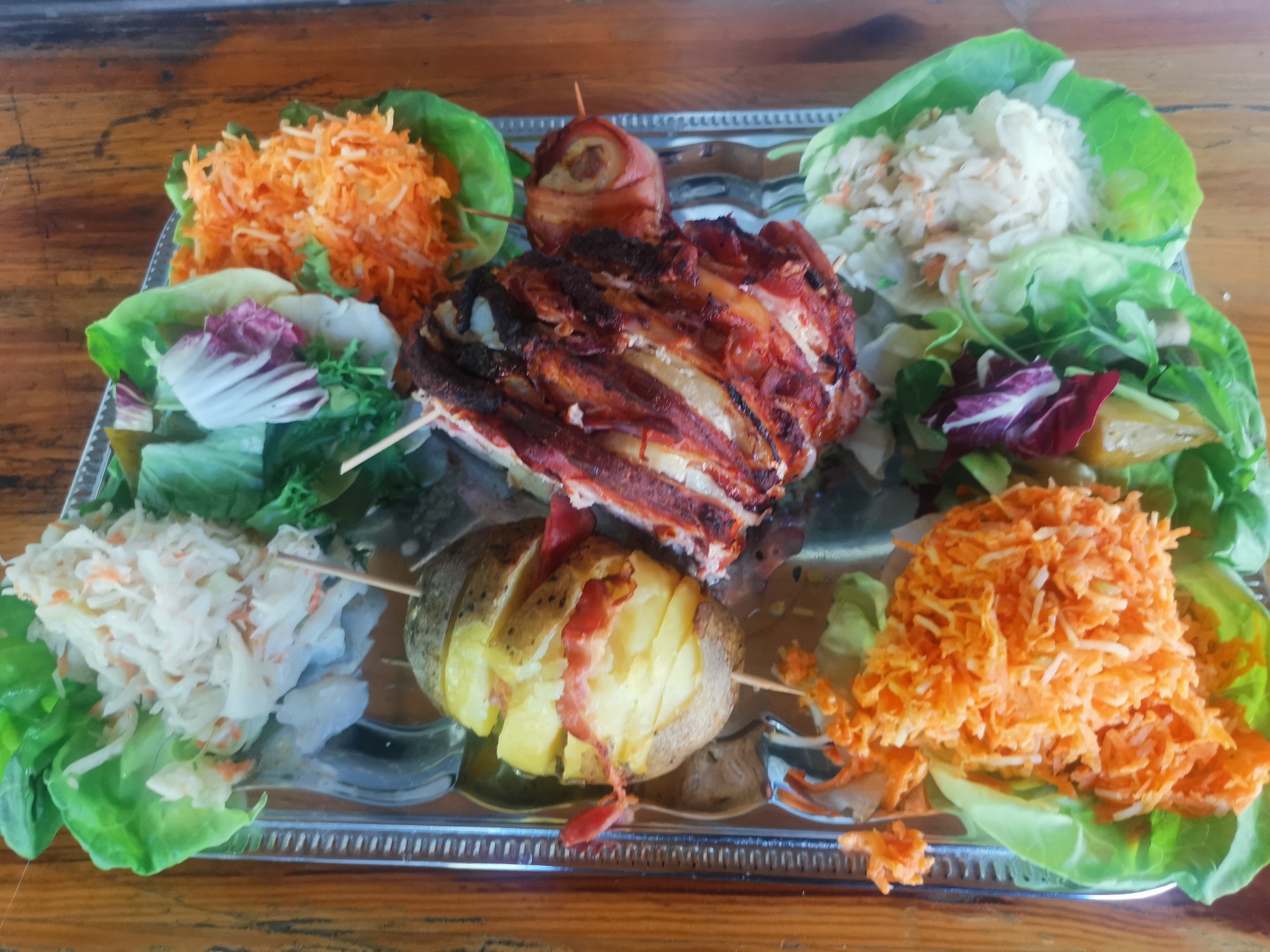 Srebrna taca z kompozycją dań: pieczone ziemniaki z boczkiem na szpadce, mięso zawijane w bekon, surówka z marchwi i kapusty, całość udekorowana liśćmi sałaty.