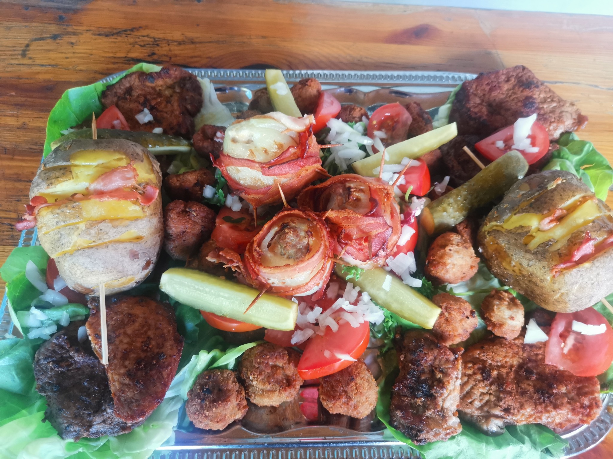 Srebrna patera z różnorodnymi przekąskami: pieczone ziemniaki z boczkiem na wykałaczkach, mięsne kulki owinięte boczkiem, kotleciki, ogórki kiszone, pomidory i posiekana cebula, wszystko...