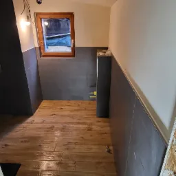 Wnętrze łazienki w trakcie wykańczania: drewnopodobne płytki podłogowe, szare płytki ścienne do połowy wysokości, fragment instalacji elektrycznej z żarówką, okno z drewnianą ramą, surowy otwór...