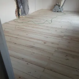 Jasna, odnowiona podłoga z desek po cyklinowaniu, z widoczną maszyną do cyklinowania i zabezpieczonym folią otworem w ścianie, w pustym pomieszczeniu.