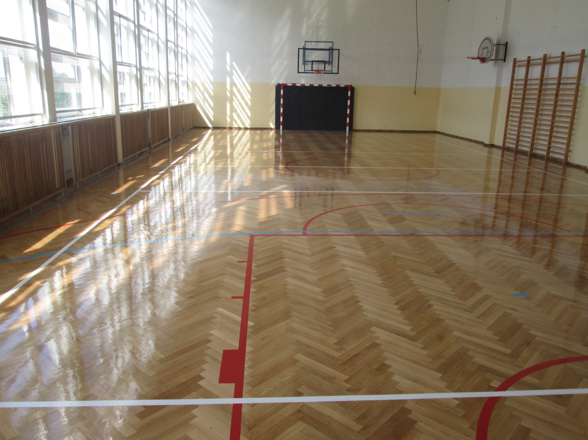 Parkiet w jodełkę na sali gimnastycznej z liniami boiska, oświetlony promieniami słońca wpadającymi przez okna. Widoczne bramki i drabinki.