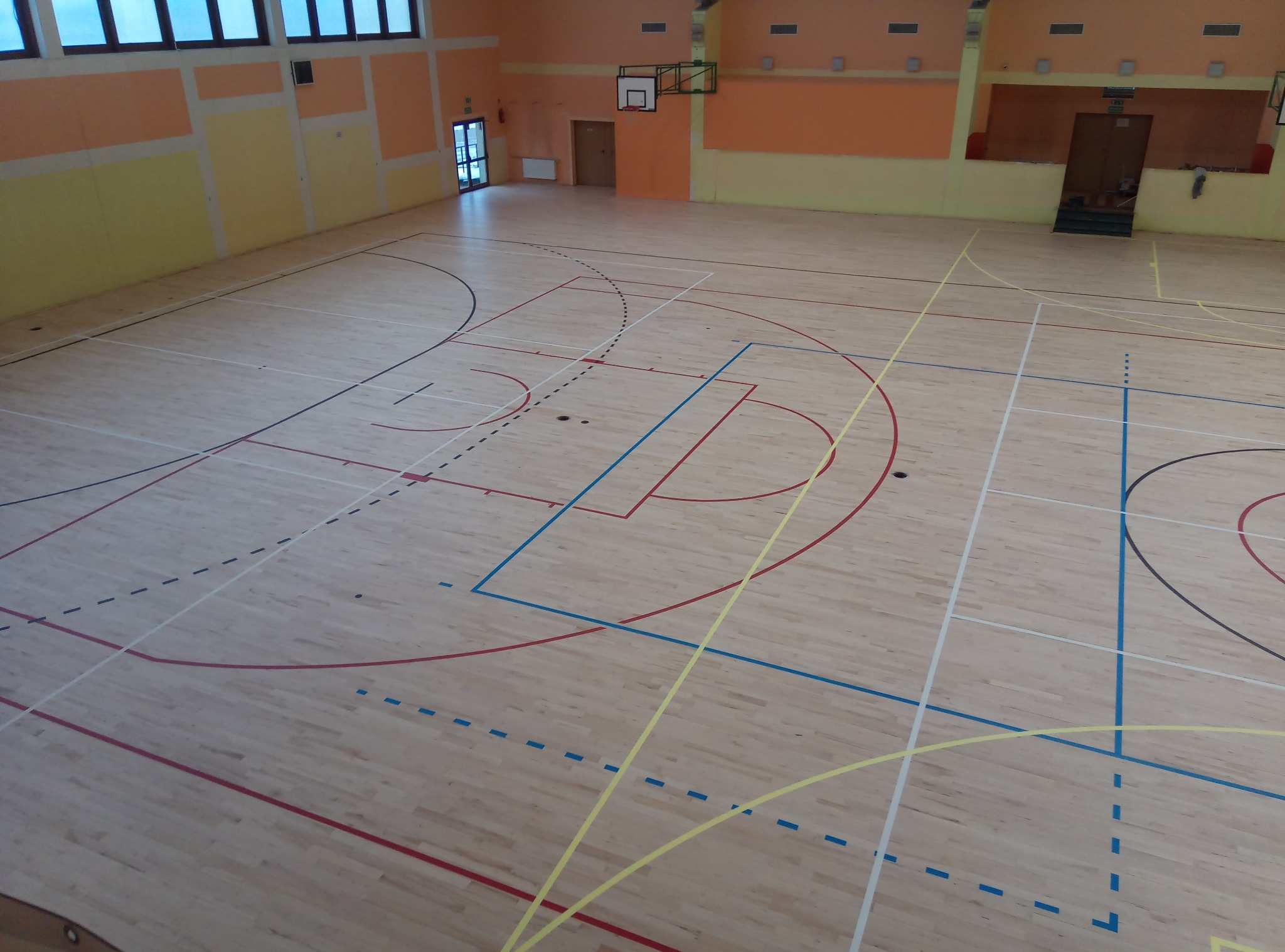 Sala gimnastyczna z parkietem w jasnym odcieniu drewna, z wyraźnie zaznaczonymi liniami boiska w kolorach: niebieskim, czerwonym, żółtym i białym. Widok z góry.