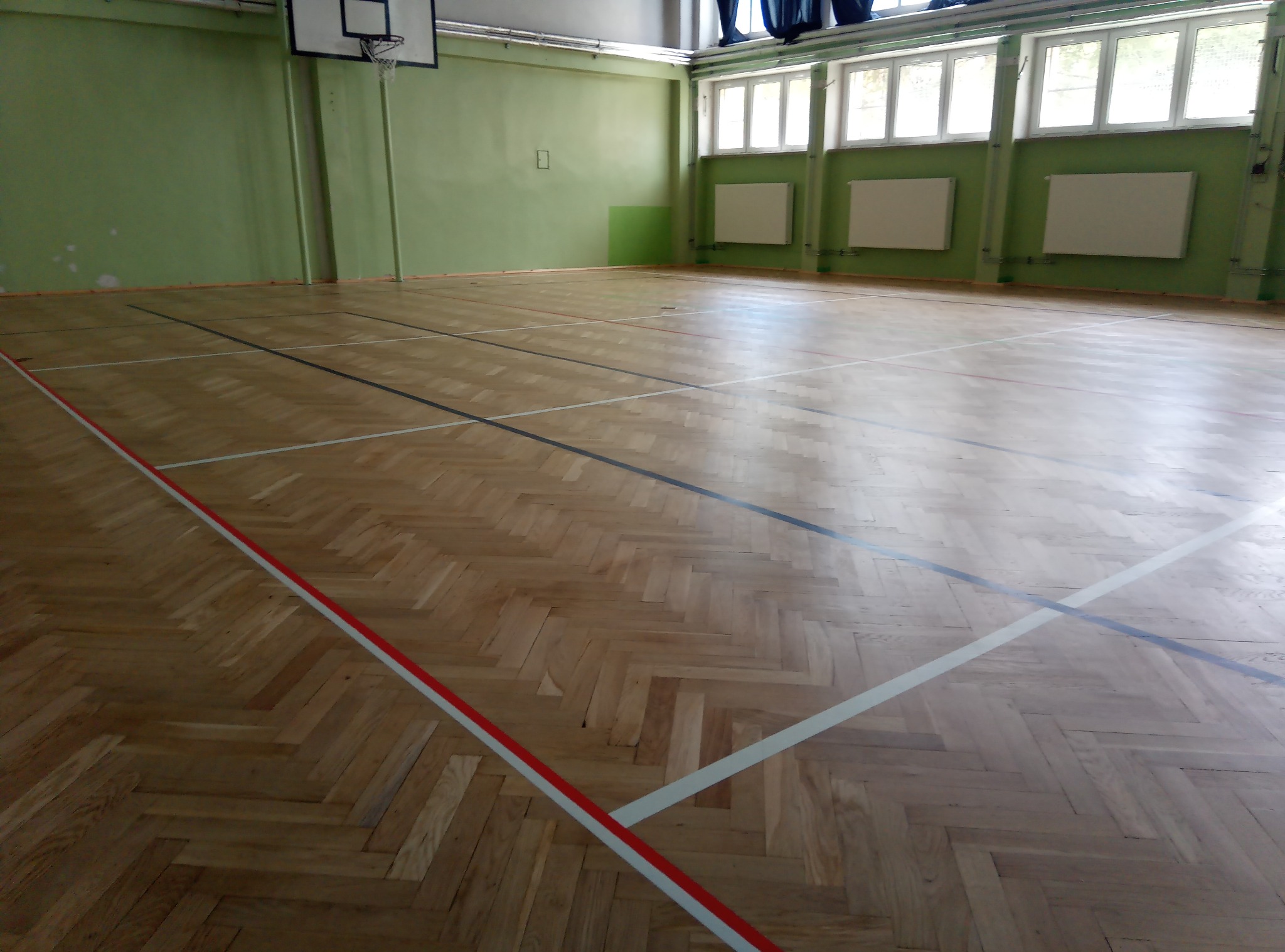 Parkiet w hali sportowej z wyraźnie zaznaczonymi liniami boiska w kolorach: czerwonym, białym i czarnym. Parkiet ułożony w jodełkę, ściany zielone.