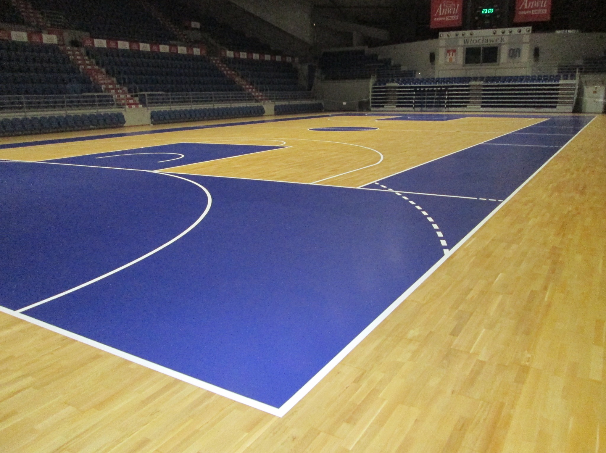 Parkiet sportowy w hali, z wyraźnie zaznaczonymi liniami boiska w kolorze białym i niebieskim. Widoczne trybuny i tablica wyników.
