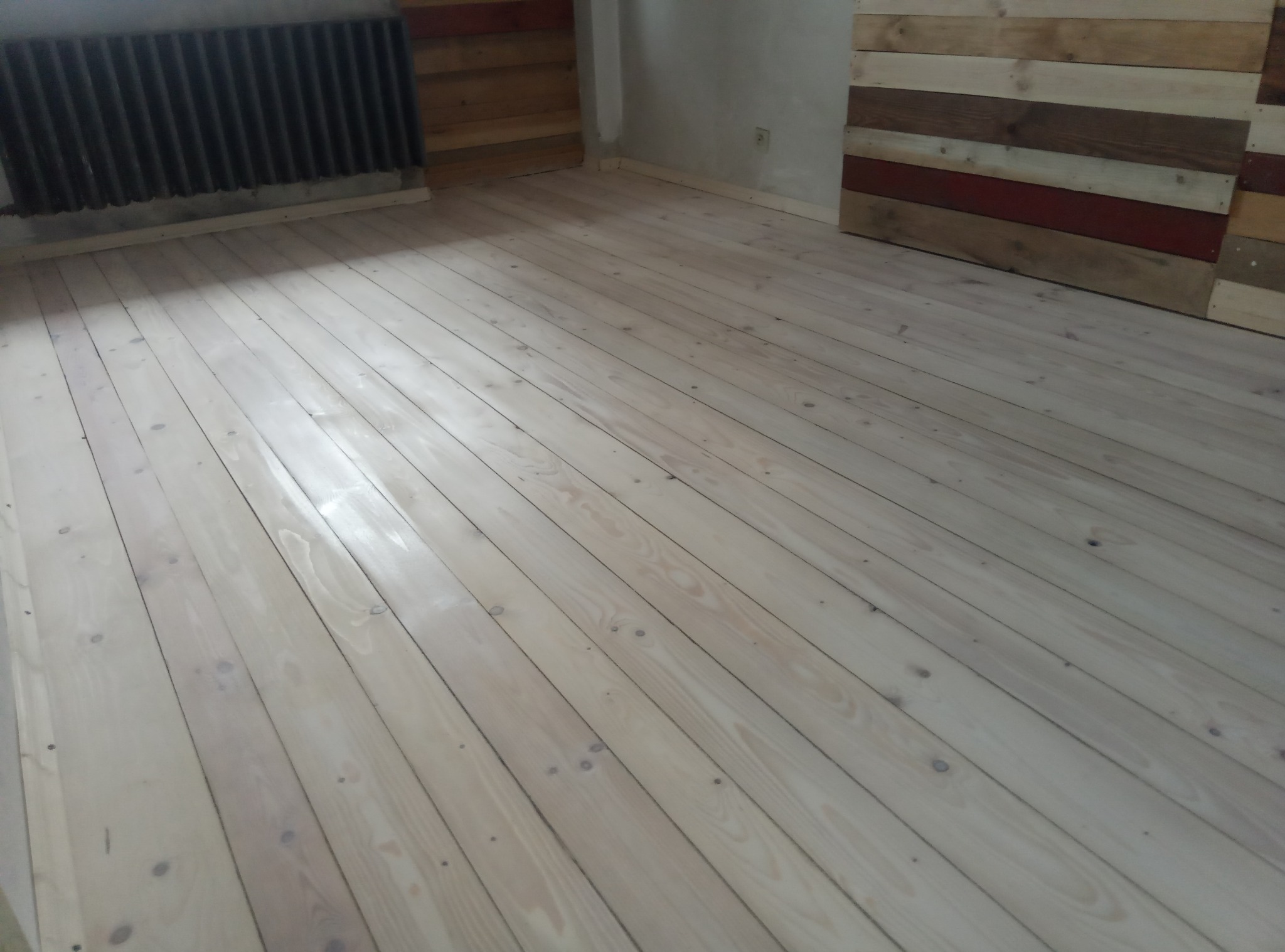 Jasna, odnowiona podłoga z desek w pokoju z drewnianą boazerią. Widoczny grzejnik. Parkiet po cyklinowaniu, przygotowany do dalszej obróbki.