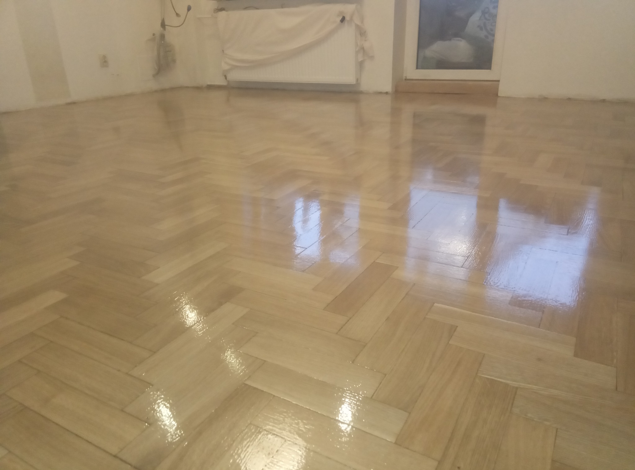 Błyszcząca, jasna podłoga z parkietu ułożonego w jodełkę, świeżo po lakierowaniu, odbija światło w pustym pomieszczeniu. Widoczny fragment okna i grzejnika.