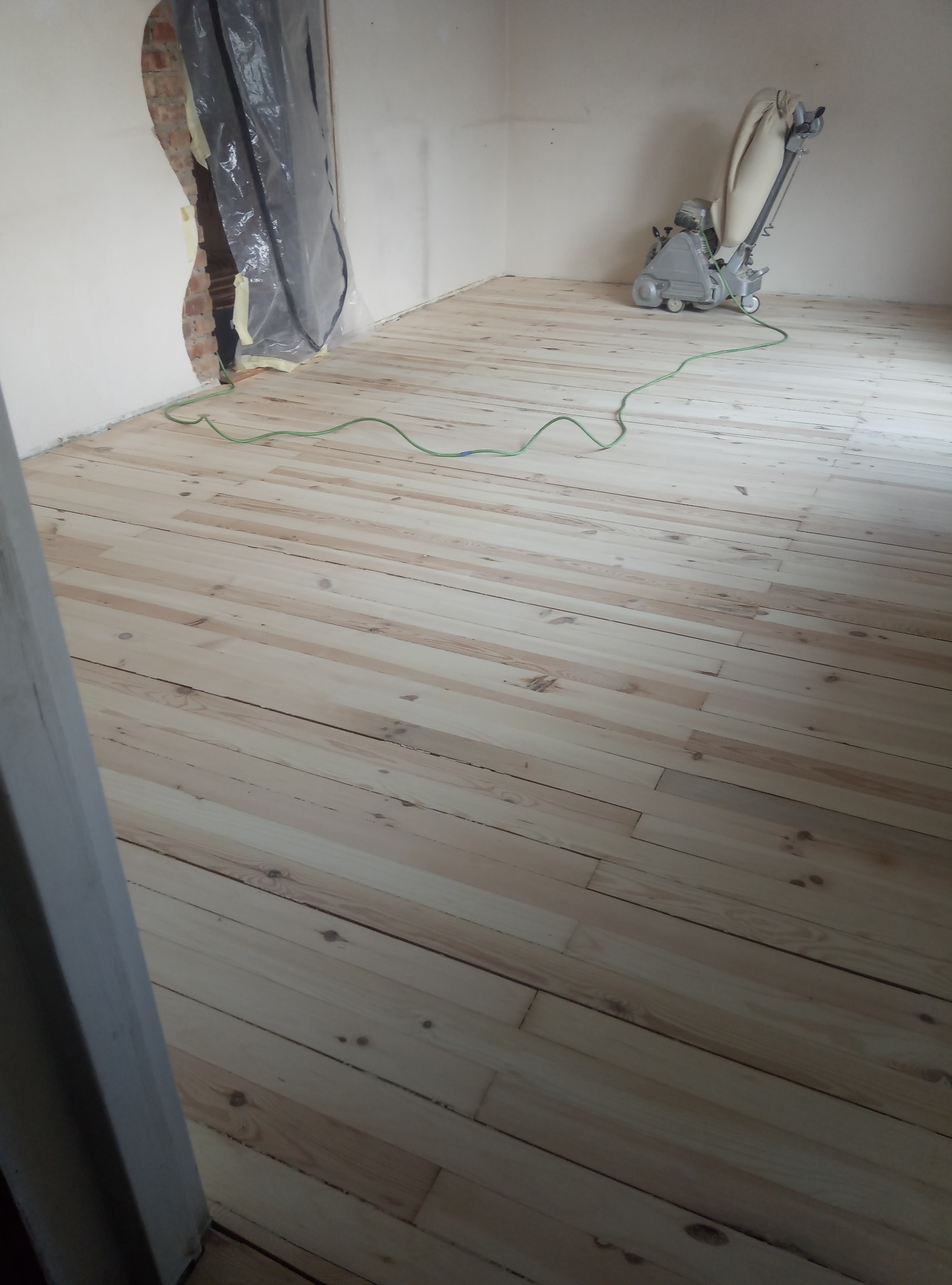 Jasna, odnowiona podłoga z desek po cyklinowaniu, z widoczną maszyną do cyklinowania i zabezpieczonym folią otworem w ścianie, w pustym pomieszczeniu.