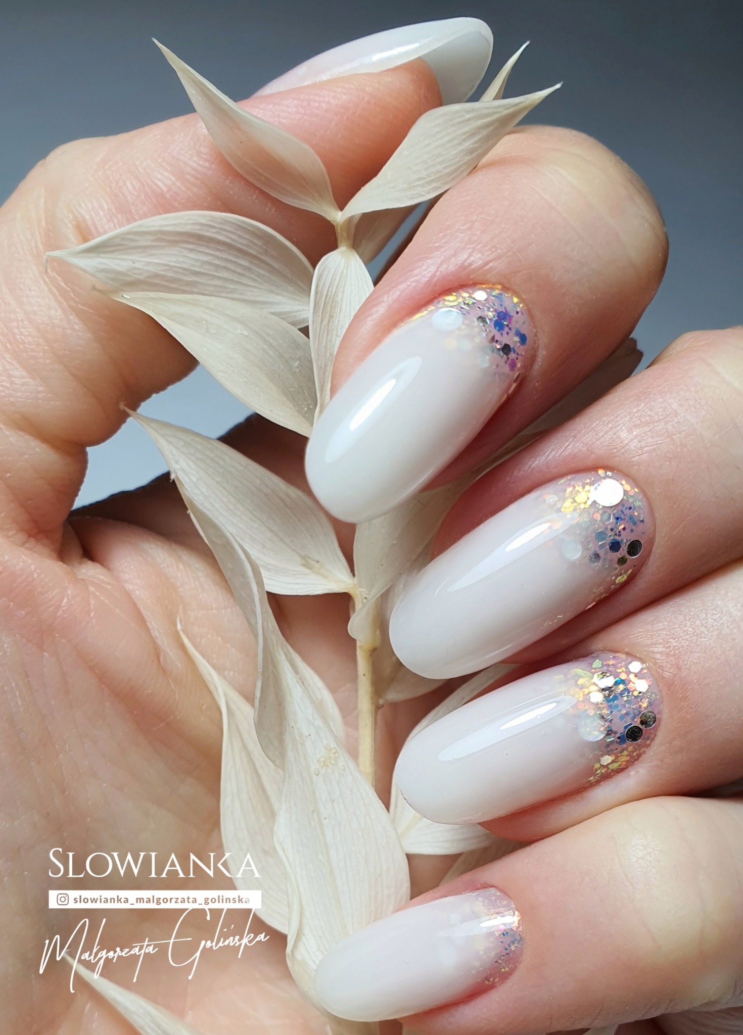 Delikatne, białe paznokcie z opalizującym brokatem na końcach, trzymane w dłoni z suchą gałązką, prezentacja stylizacji.