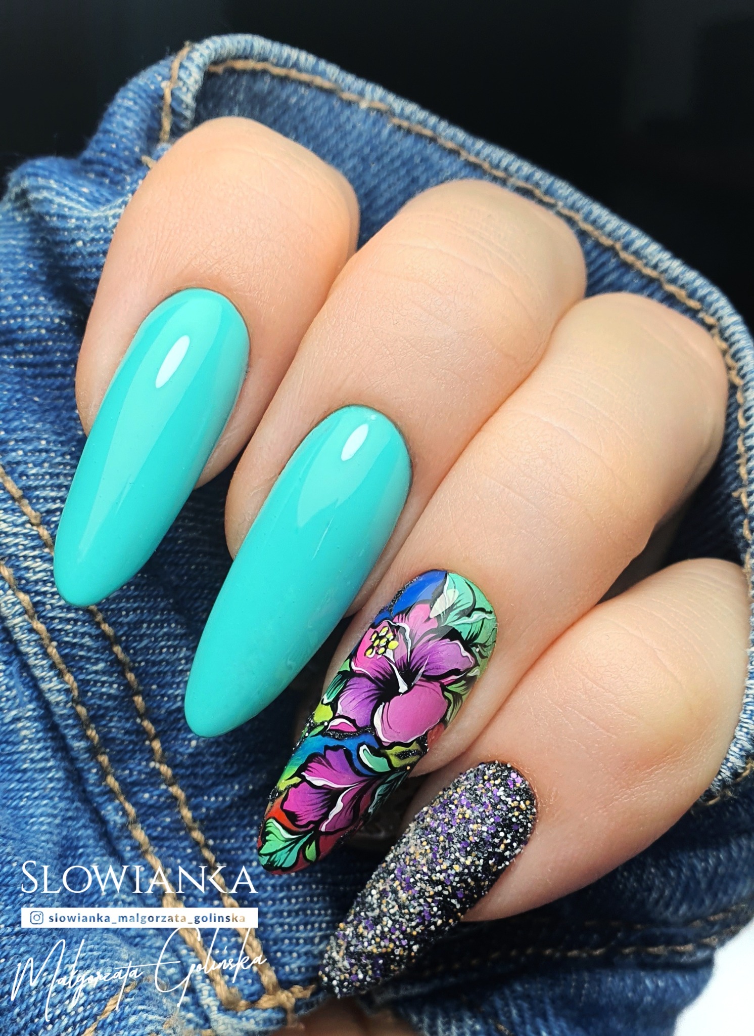 Dłoń z turkusowymi, błyszczącymi i kwiatowymi stylizacjami paznokci na tle dżinsowej tkaniny.