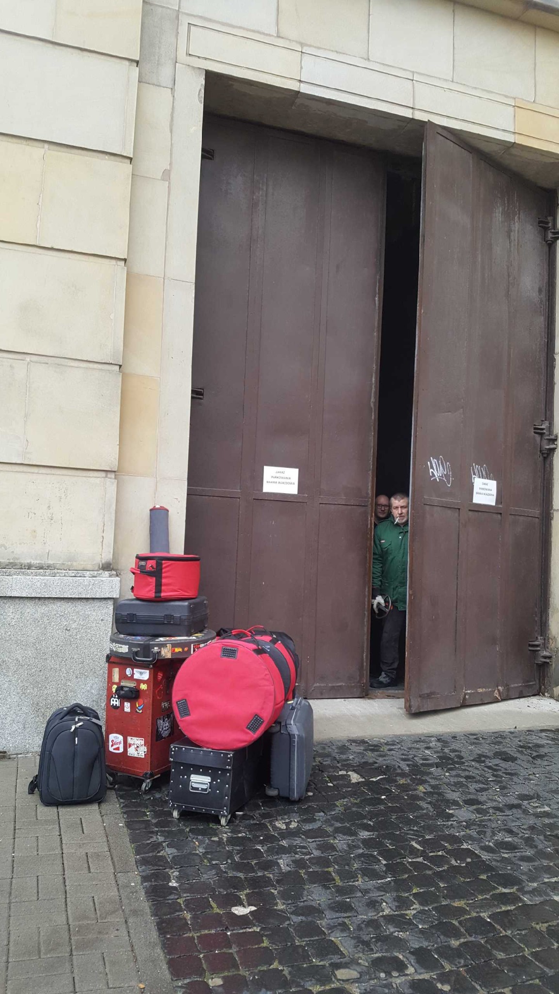 Stos walizek i toreb, w tym czerwona okrągła torba, przed otwartą, brązową bramą wjazdową do budynku z kamienną elewacją; w tle widoczni dwaj mężczyźni w roboczych ubraniach.