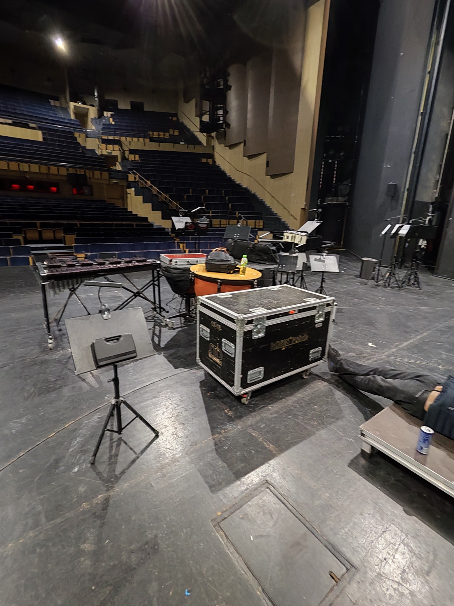 Sprzęt muzyczny (marimba, perkusja) w skrzyniach transportowych na scenie teatru przed widownią, z widocznymi nogami osoby leżącej obok.