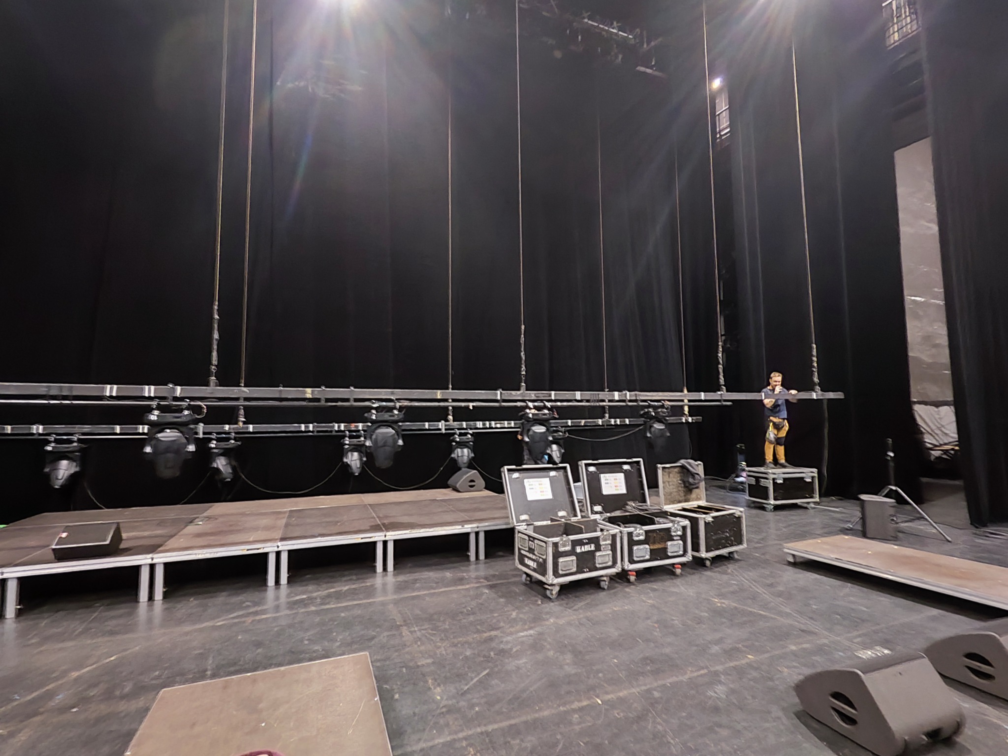 Przygotowanie sceny teatralnej: mężczyzna w szelkach stoi na skrzyni, montując oświetlenie na czarnym tle zasłon, widoczne skrzynie z kablami i głośniki na podłodze sceny.