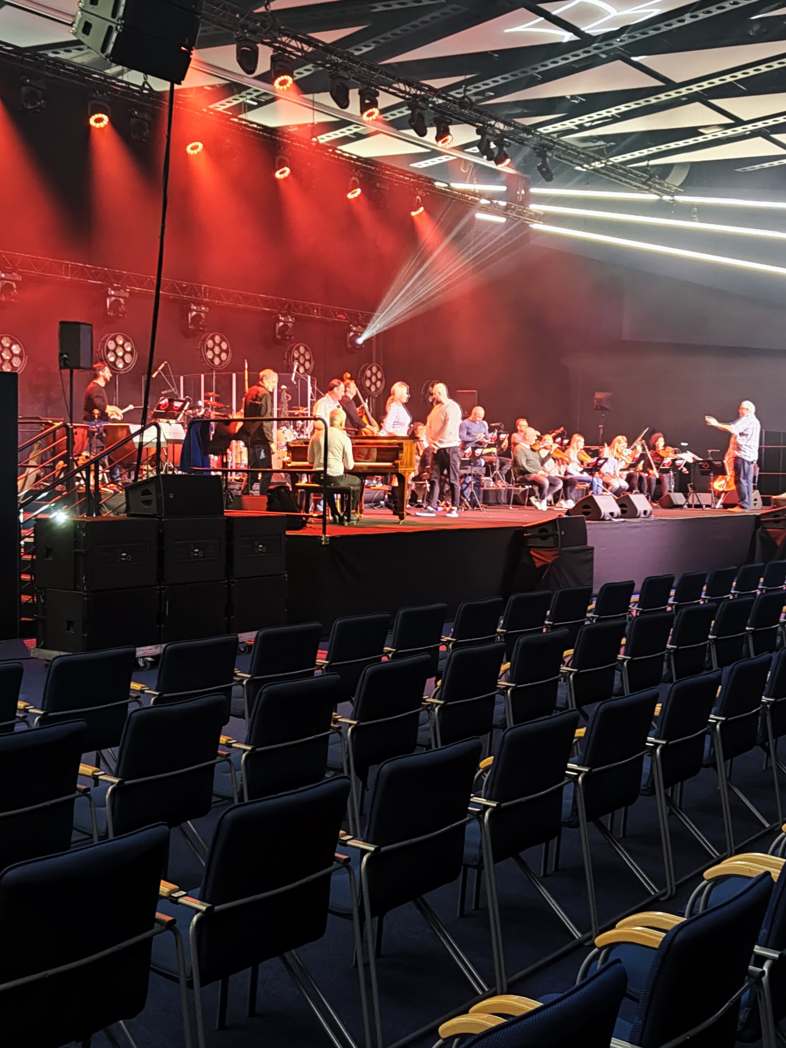 Zespół muzyczny na scenie oświetlonej czerwonymi reflektorami, widok z pustej widowni z rzędami krzeseł.