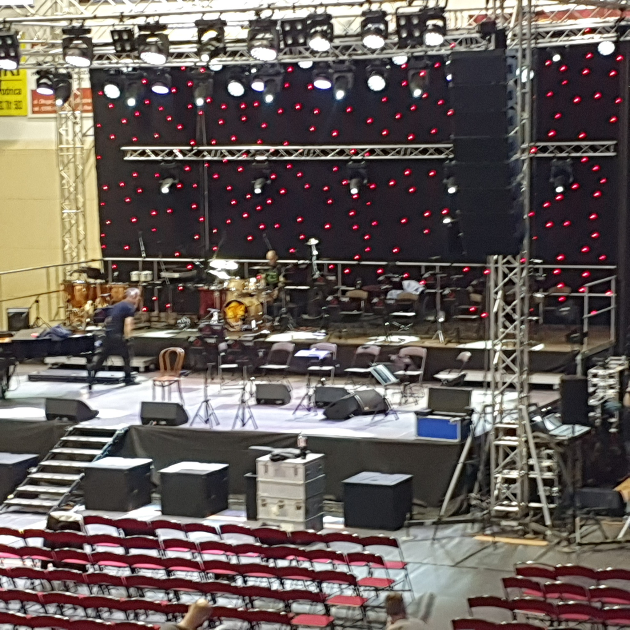 Przygotowanie sceny: mężczyzna ustawia sprzęt muzyczny na scenie z dekoracją z czerwonych światełek, w tle widoczne instrumenty perkusyjne, krzesła i schody na podest.