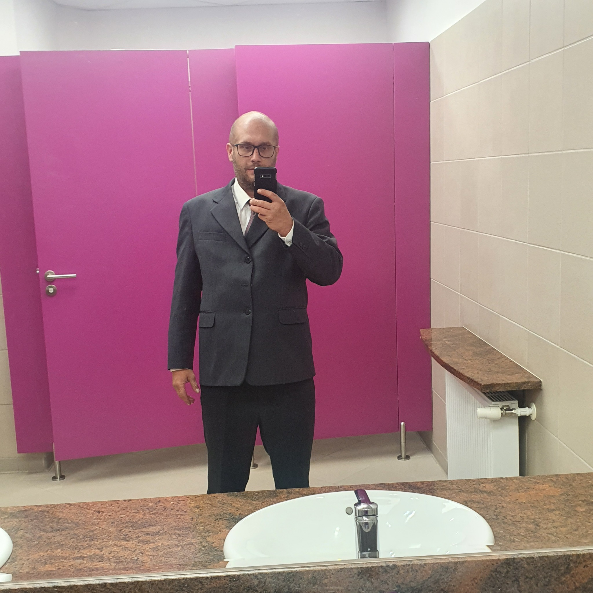Mężczyzna w garniturze robi selfie w toalecie z różowymi kabinami i umywalką z brązowym blatem.